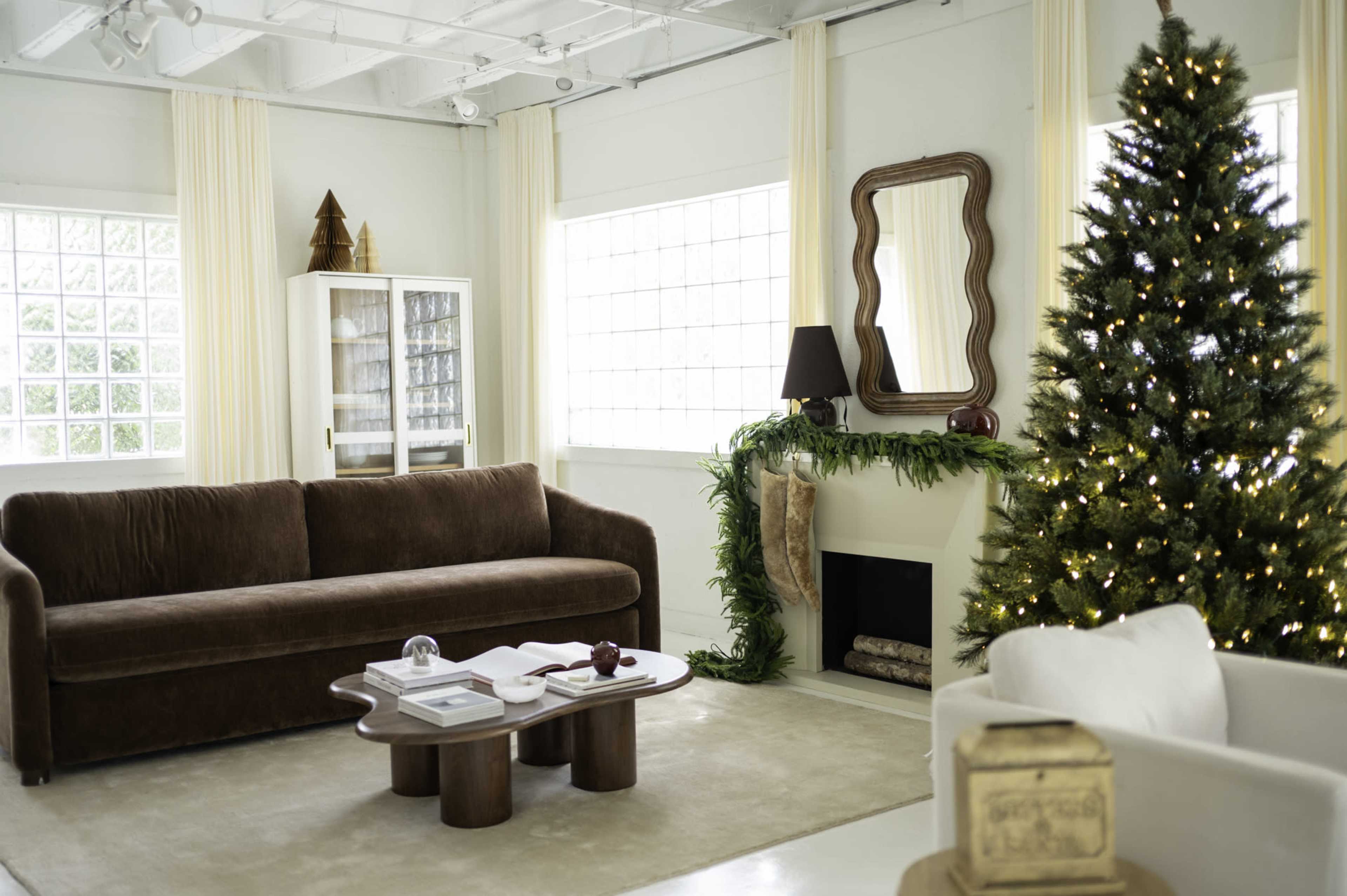 The image shows a styled living room featuring a brown couch, a white armchair, a Christmas tree decorated with lights, and a coffee table in front of a fireplace with greenery.