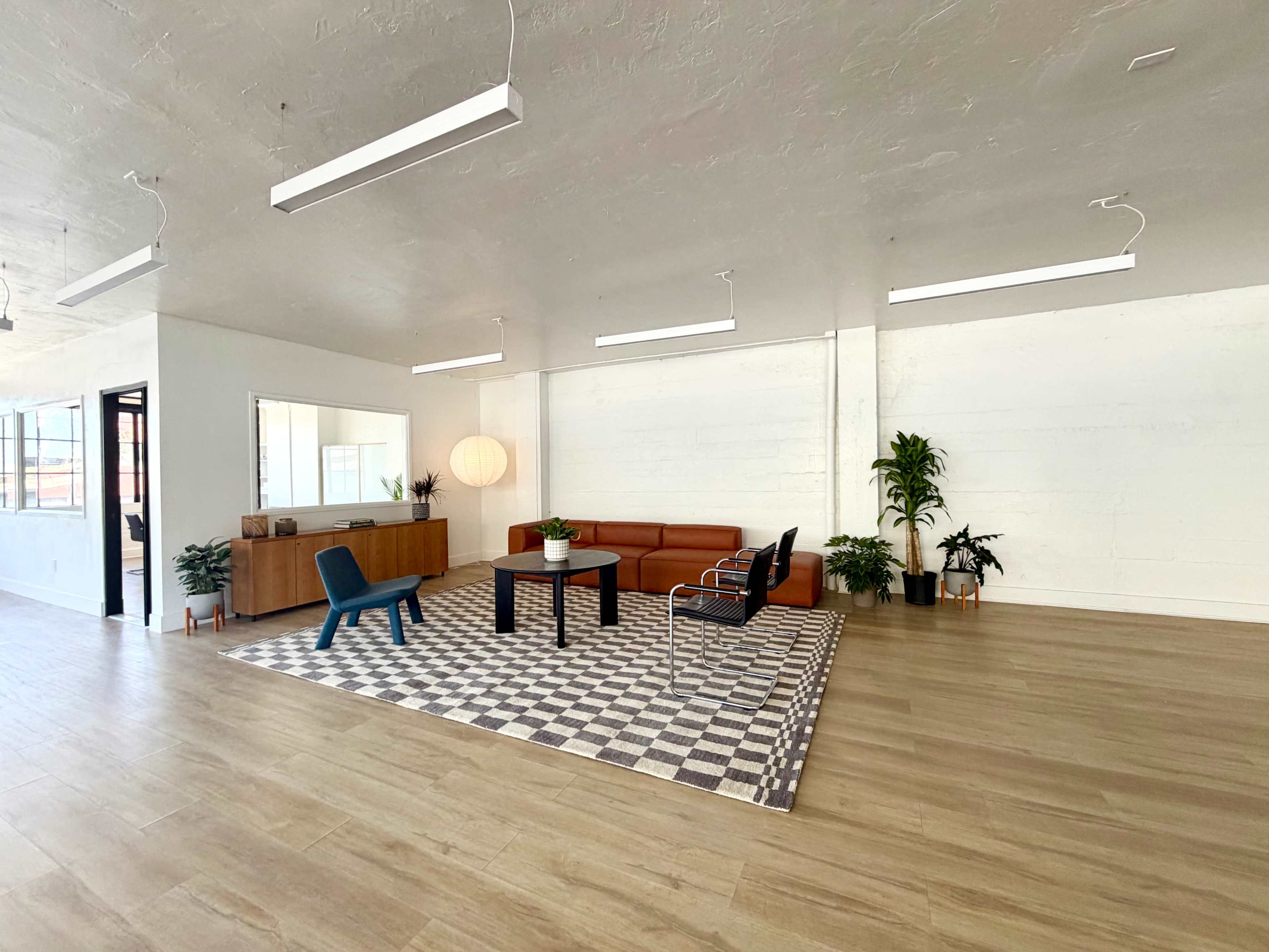The image shows a spacious, modern interior with a brown sofa, blue chair, coffee table, and plants arranged on a checkered rug.