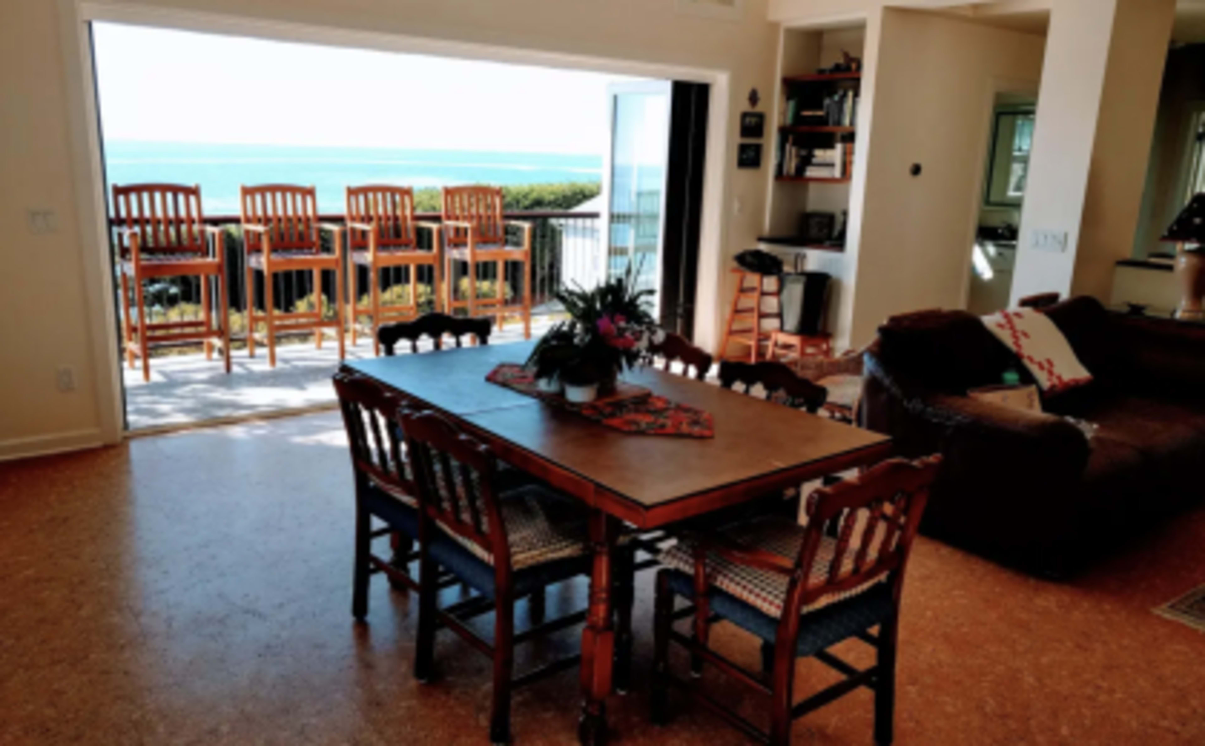 A dining table with eight chairs is positioned indoors overlooking a patio with bar stools and a view of the water.