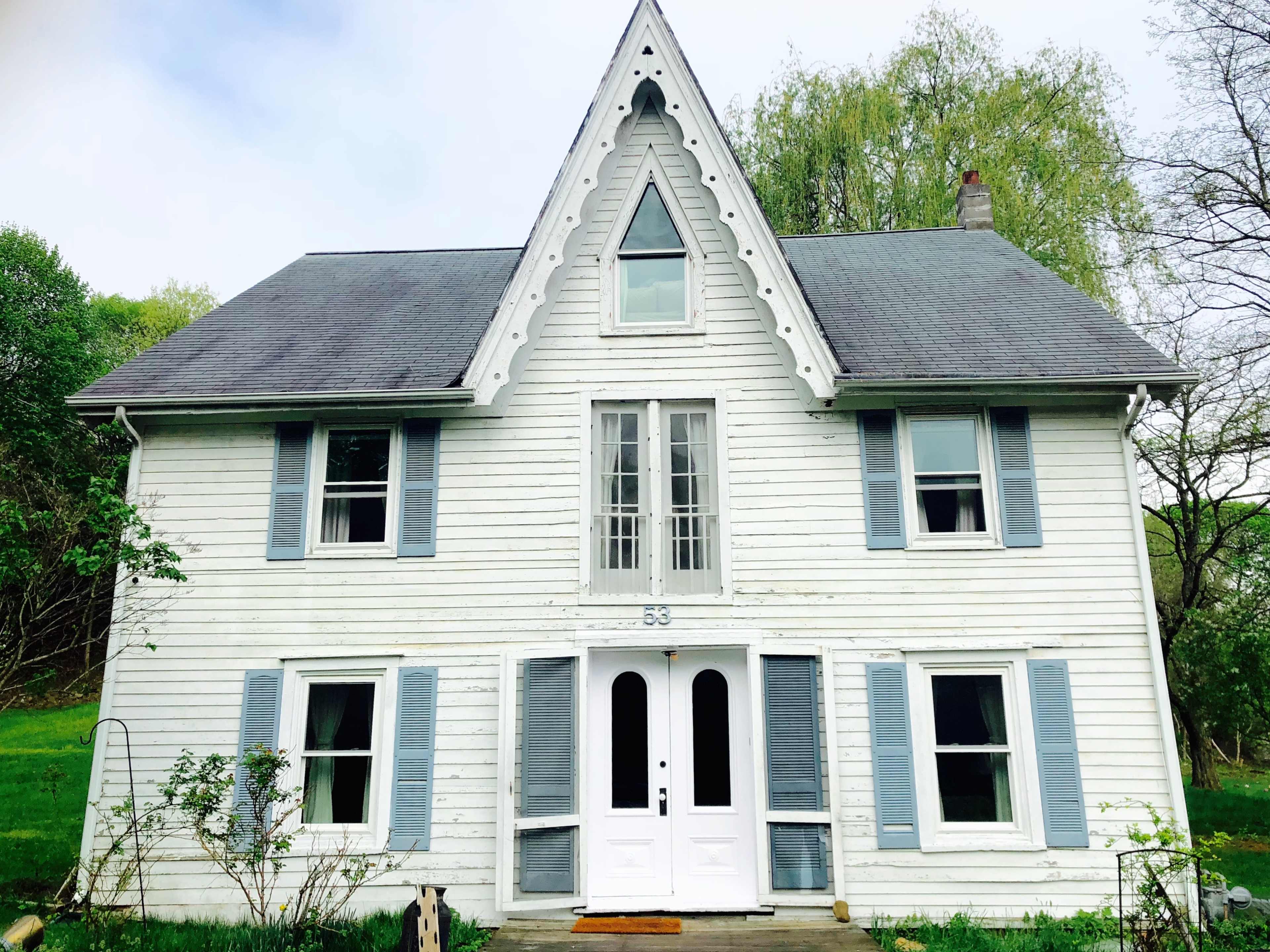 A two-story white house with a steeply pitched roof and blue shutters sits surrounded by greenery.