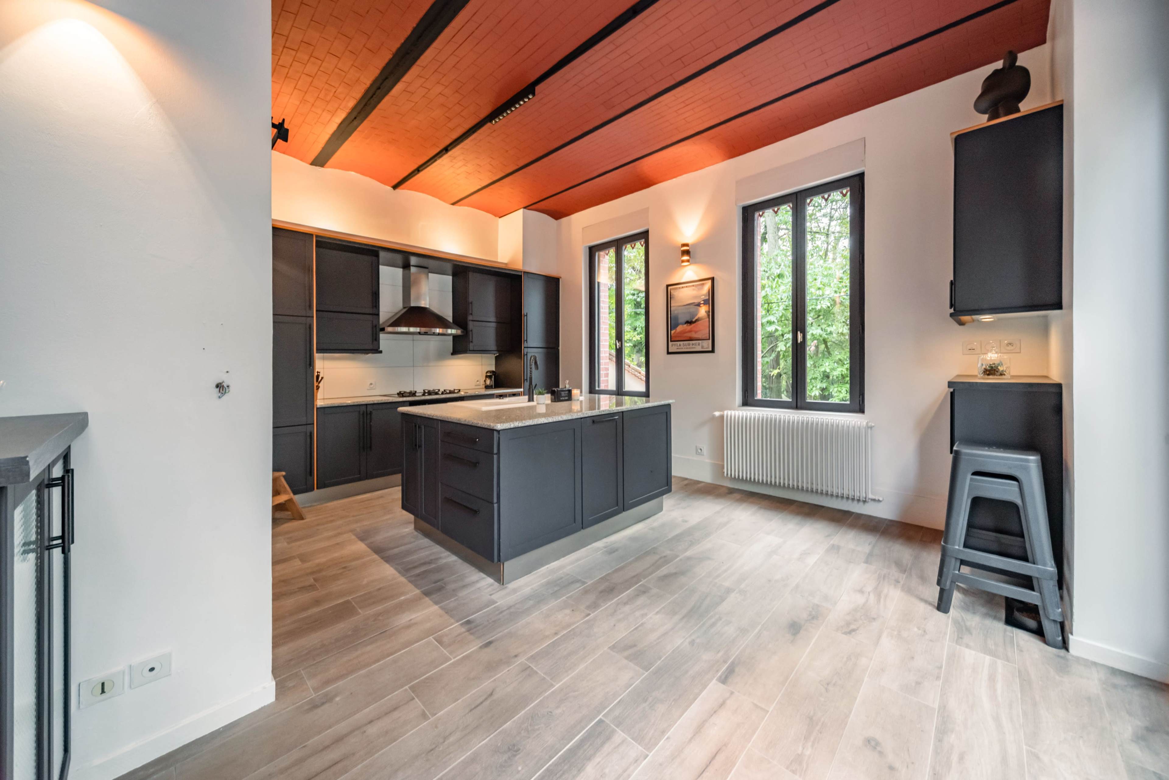 A modern kitchen features dark cabinetry, an island, and large windows that provide natural light.