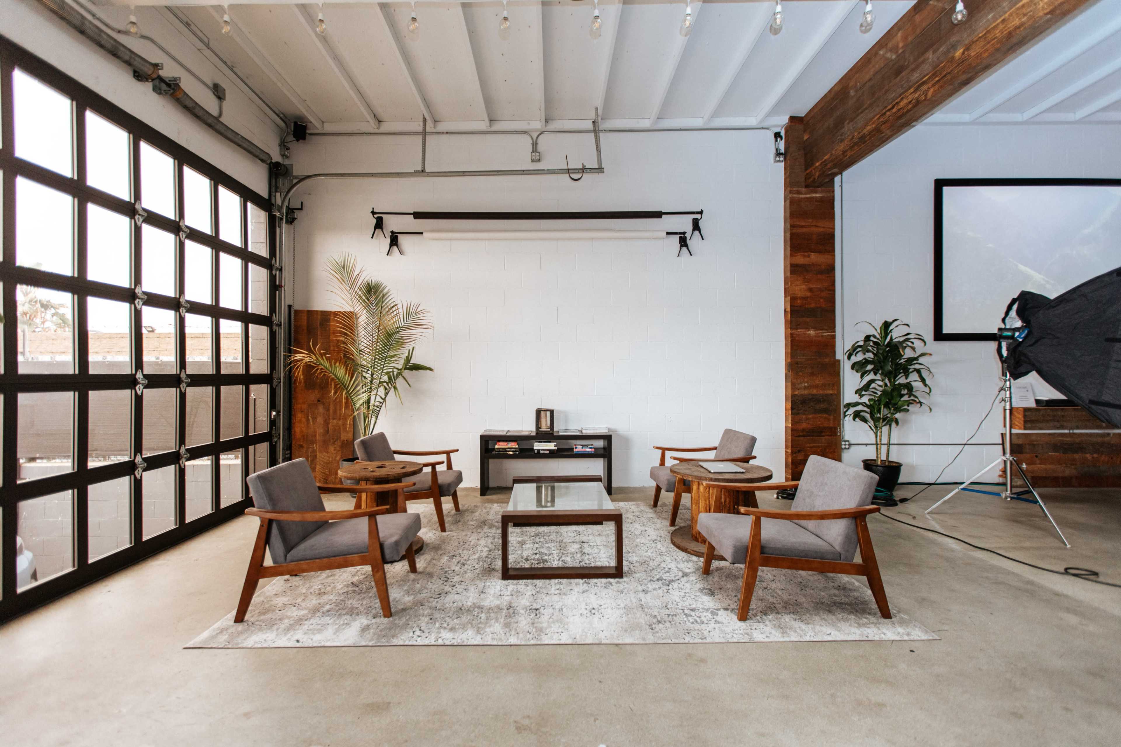A modern interior space features a seating arrangement of four wooden chairs and a glass-top coffee table on a rug, against a backdrop of large windows and greenery.