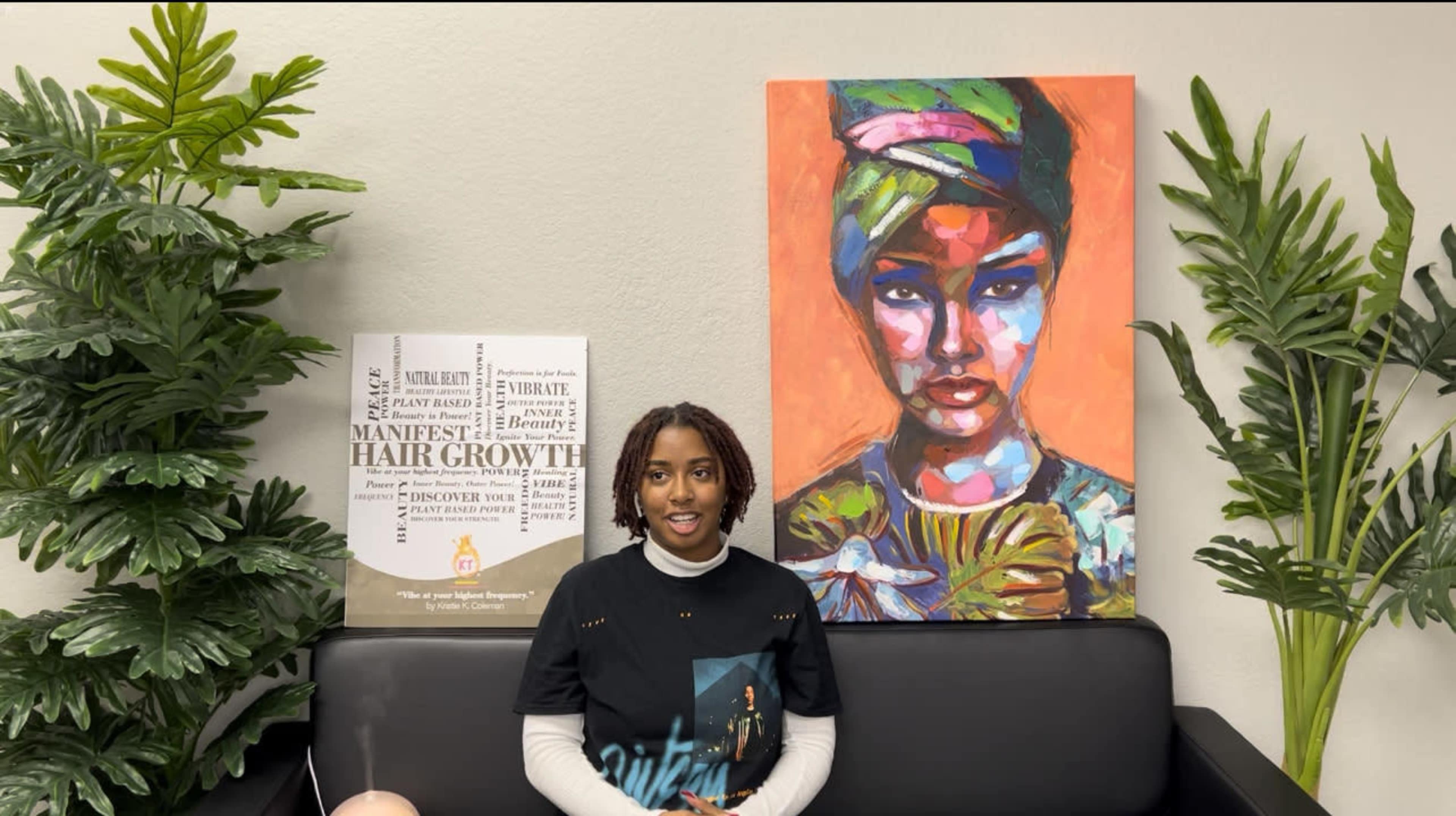 A person sits on a black couch in front of a colorful portrait and a poster related to hair growth, surrounded by indoor plants.