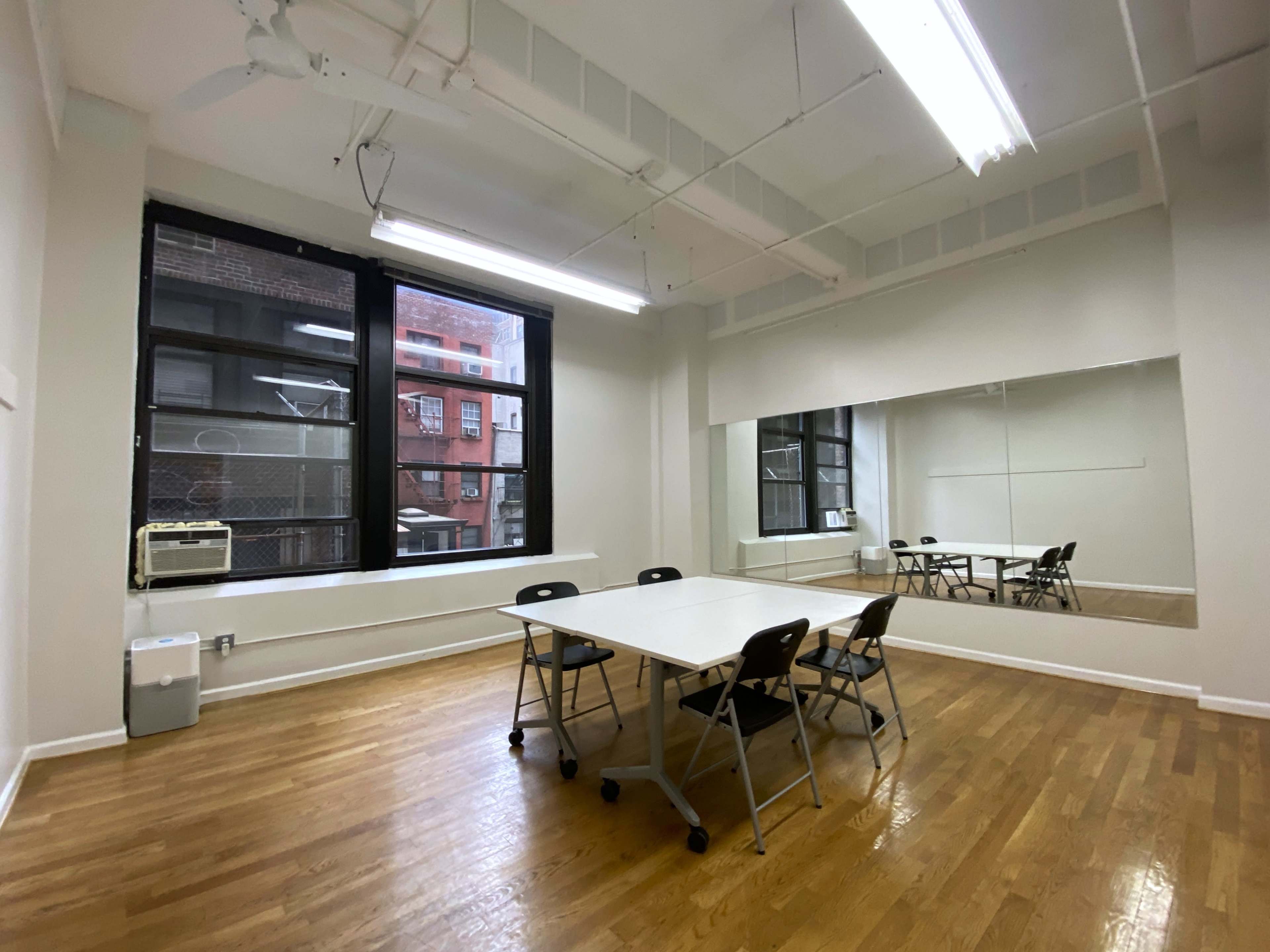 A blank, well-lit room with wooden floors, a large mirror on one wall, and a table surrounded by chairs.