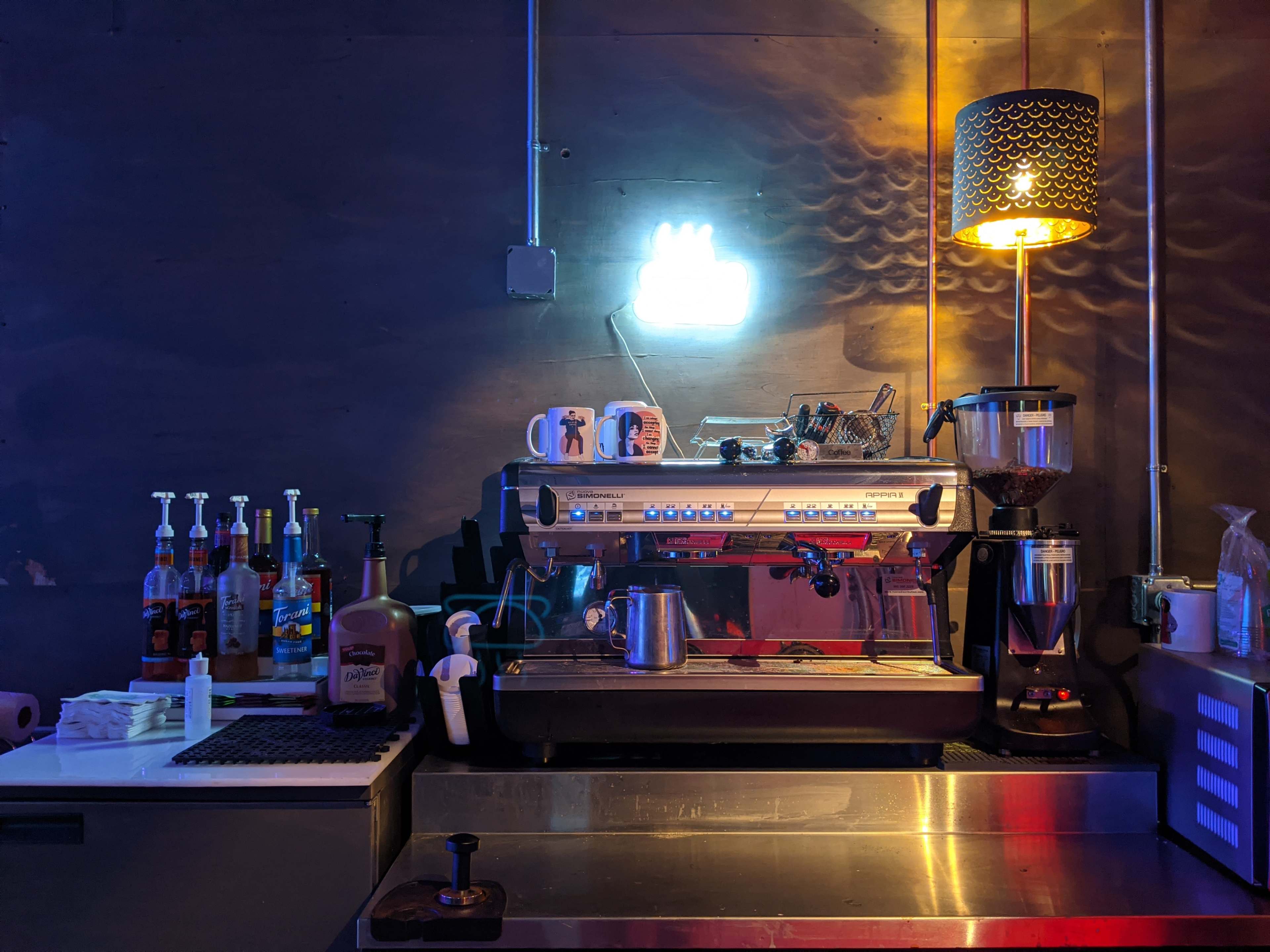 A coffee machine sits on a counter beside various bottles and tools, illuminated by a neon hand sign and a lamp.