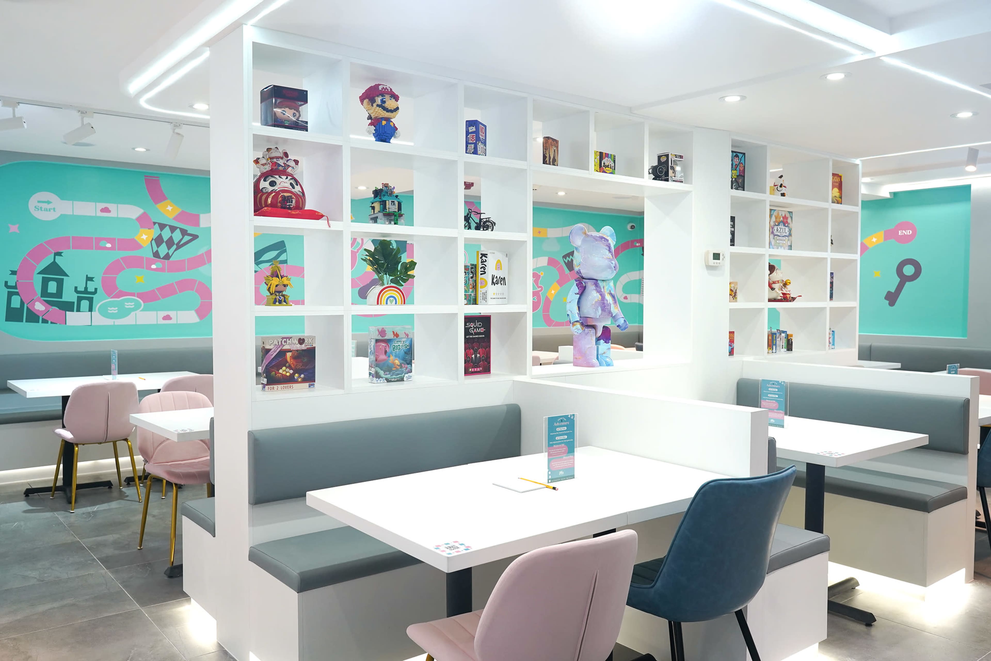 The image shows a modern cafe interior featuring a white shelving unit filled with various collectibles, colorful wall murals, and light-colored seating areas.