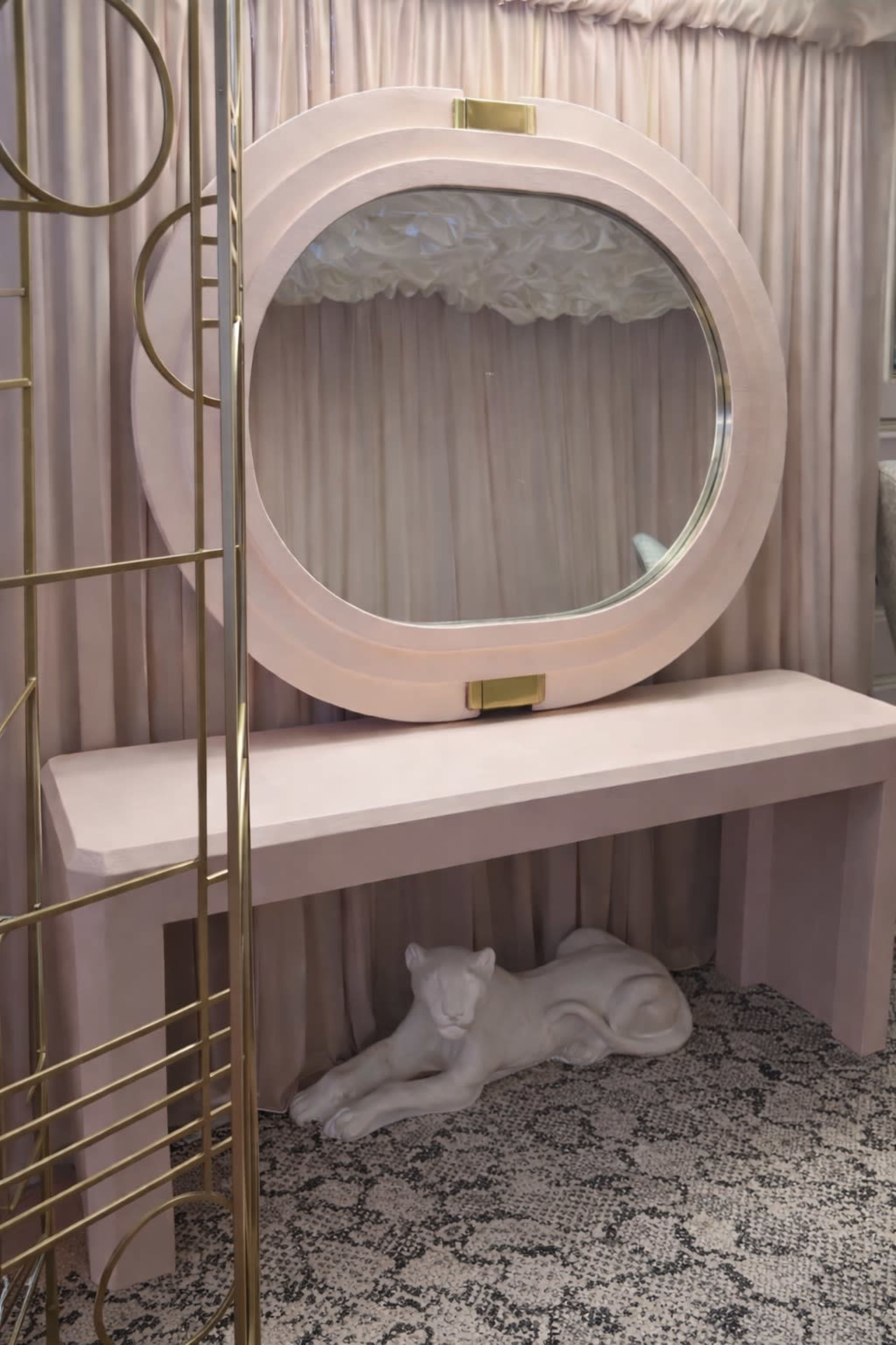 A large, oval mirror is mounted above a pale pink console table, accompanied by a white sculpted panther and set against a backdrop of soft curtains.