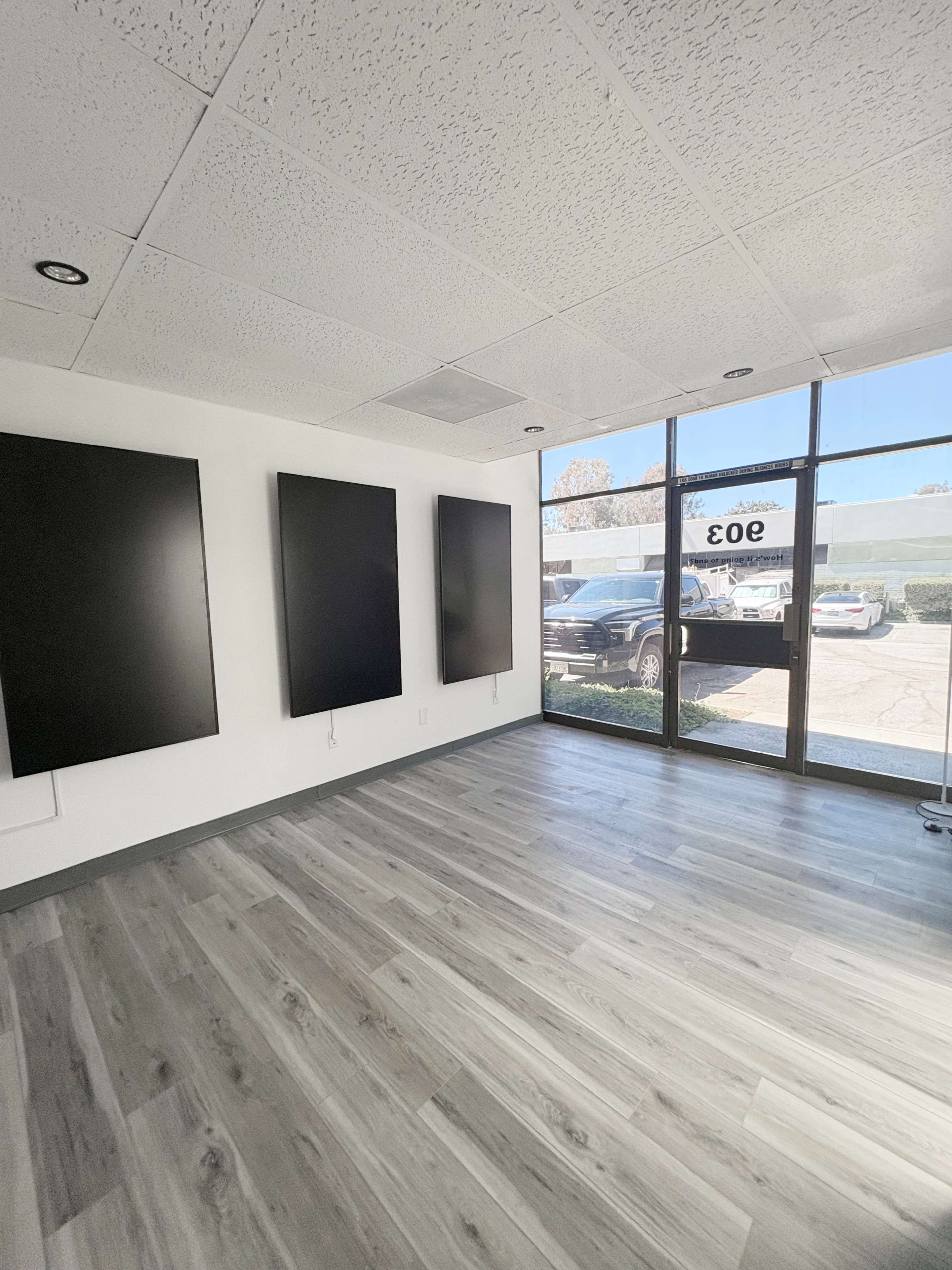 The image shows an empty room with four blank, wall-mounted panels and large windows facing an outdoor area.