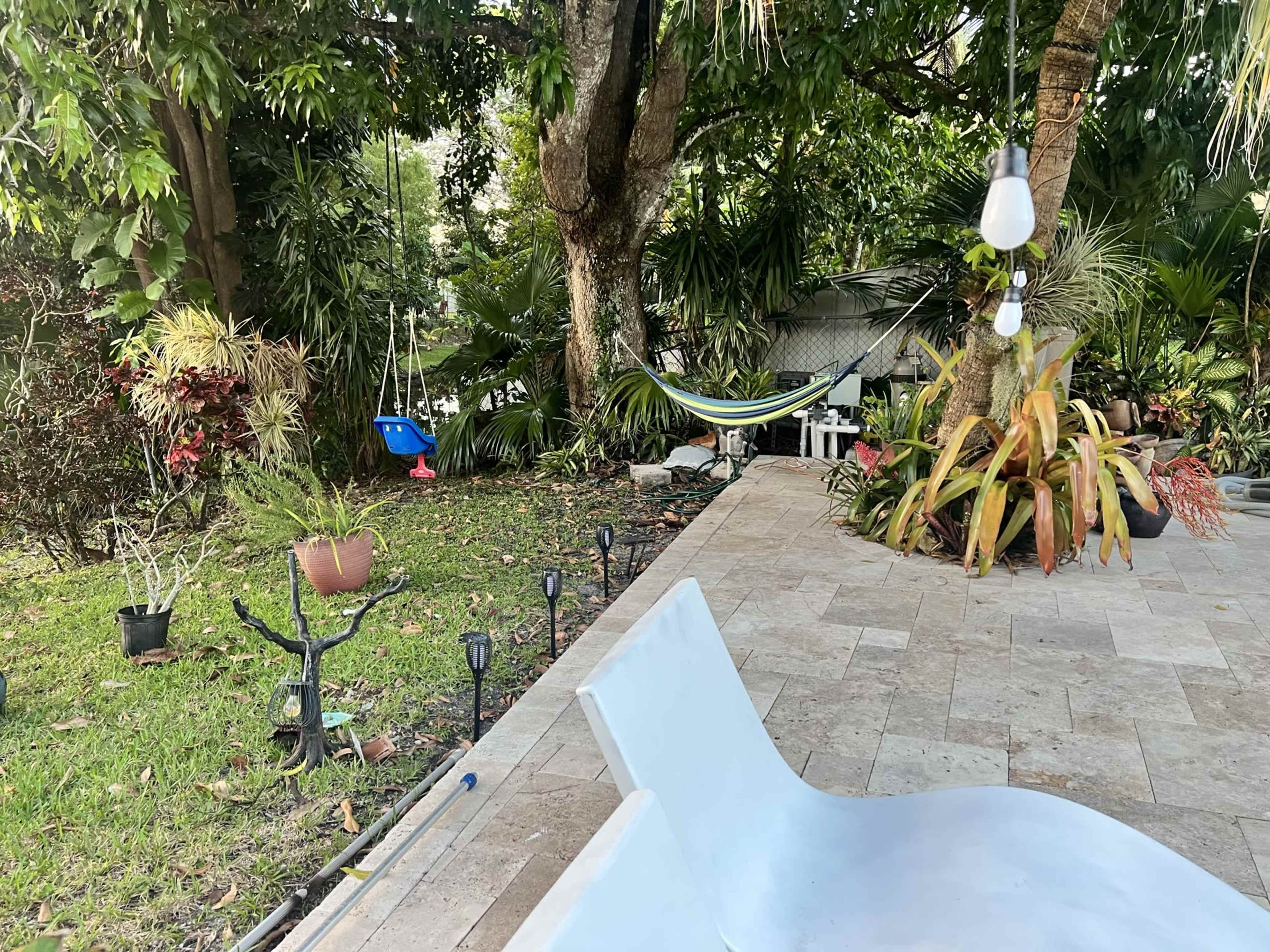 The scene shows a backyard with a hammock, a blue swing, scattered plants, and outdoor lighting along a stone patio.