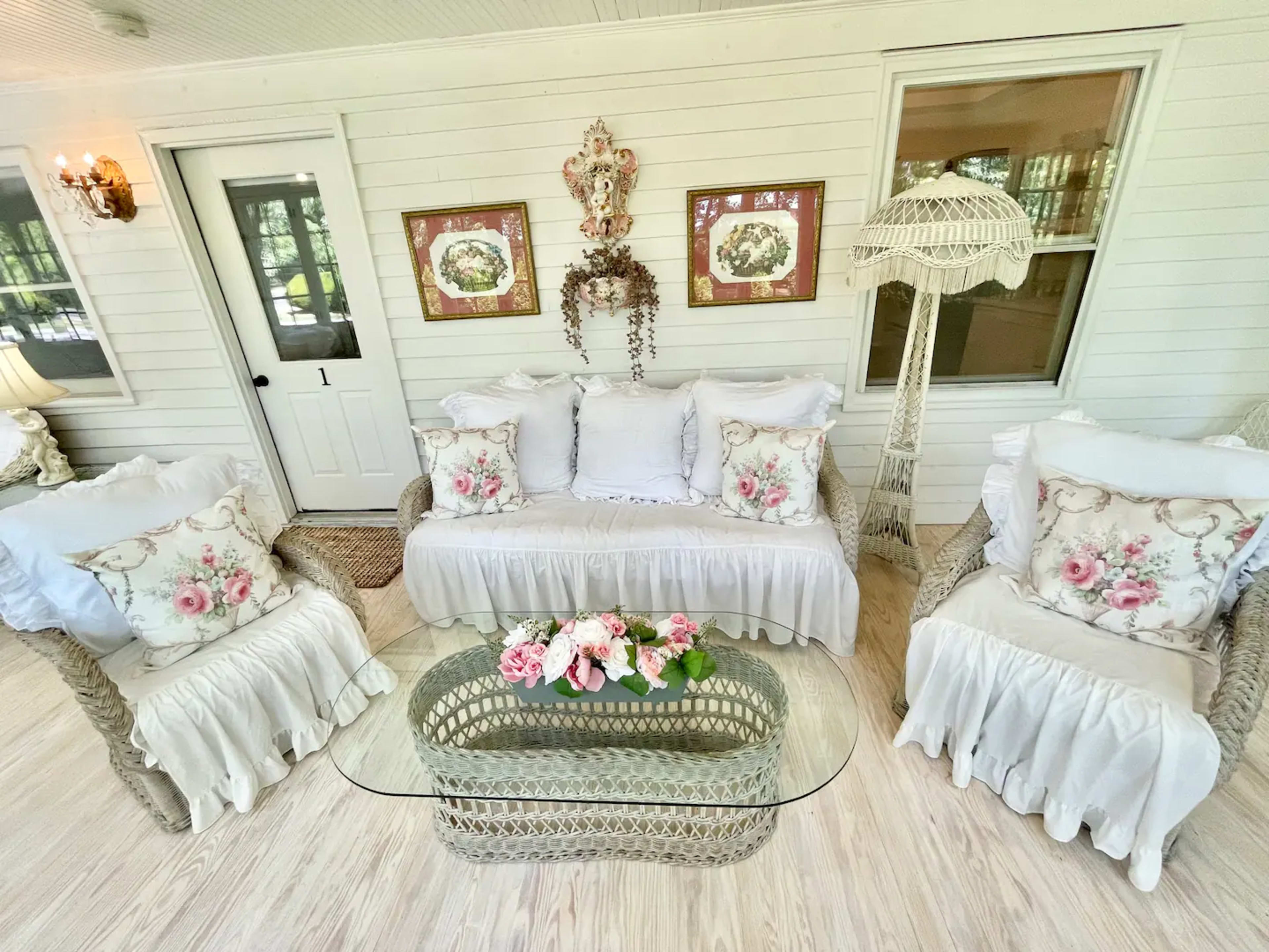 A light-filled sunroom featuring a rattan sofa with white floral pillows and a glass coffee table adorned with a floral arrangement.