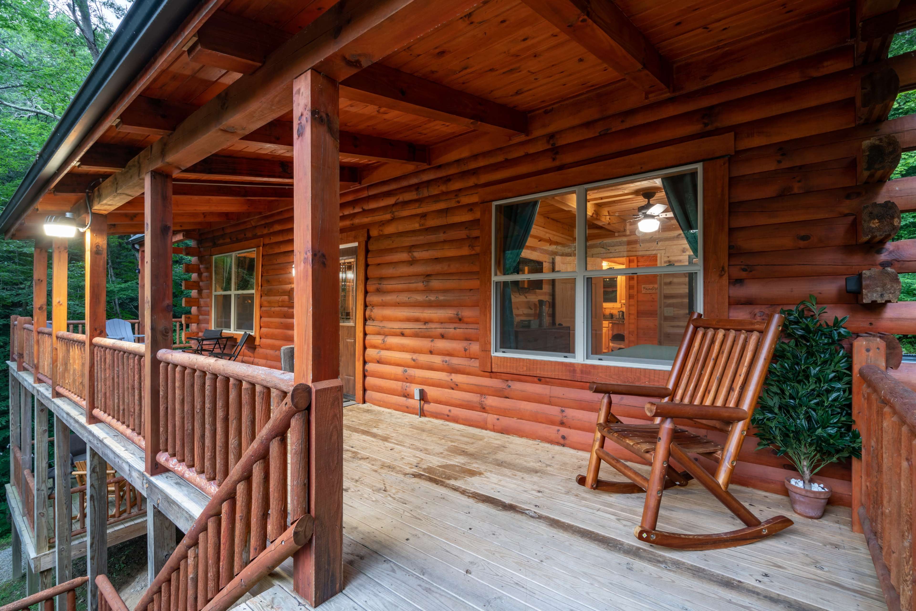 A wooden cabin with a spacious porch, featuring rocking chairs and large windows that overlook a forested area.