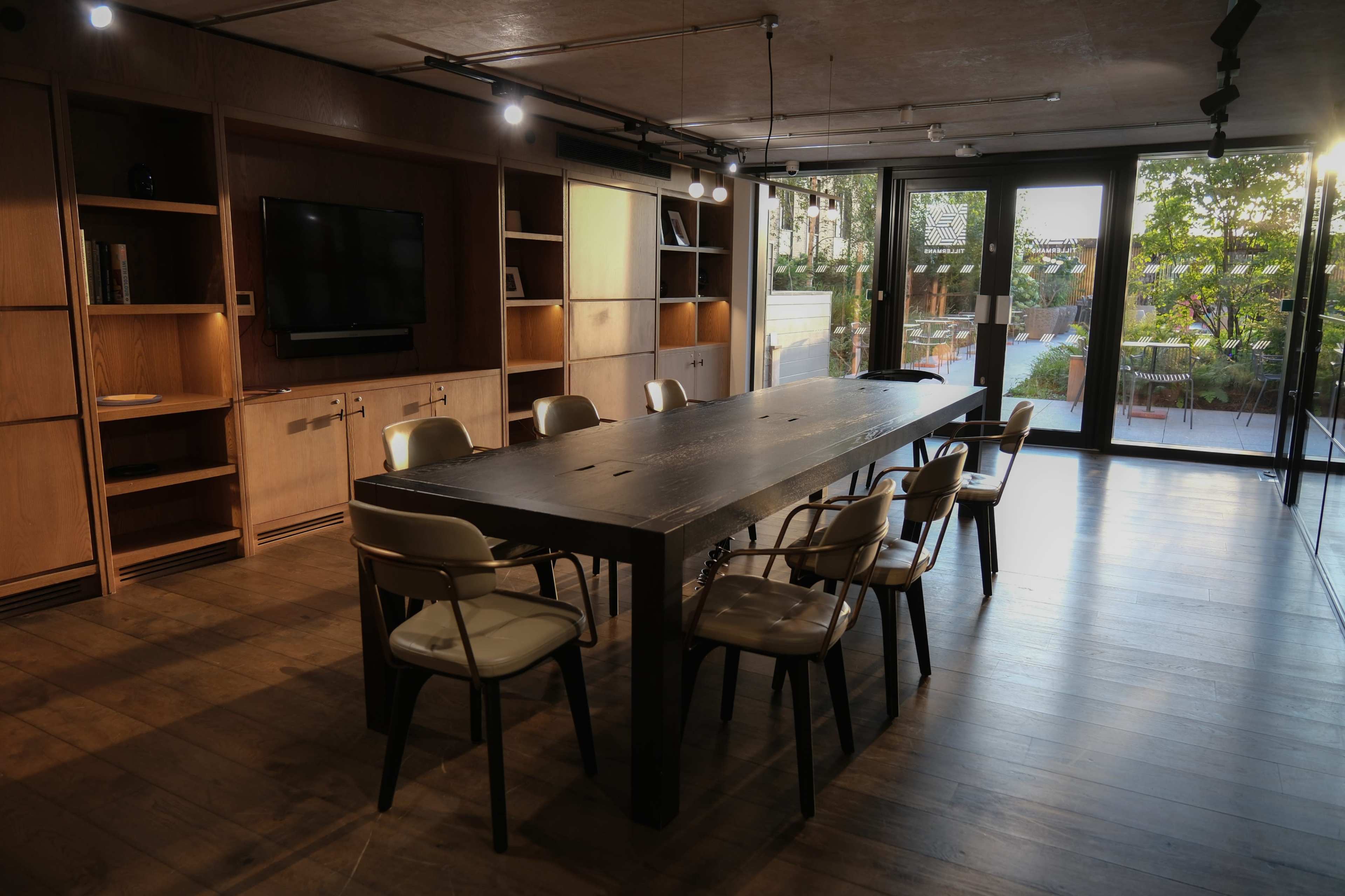 A large, wooden conference table surrounded by chairs sits in a well-lit meeting room with floor-to-ceiling windows overlooking a garden.