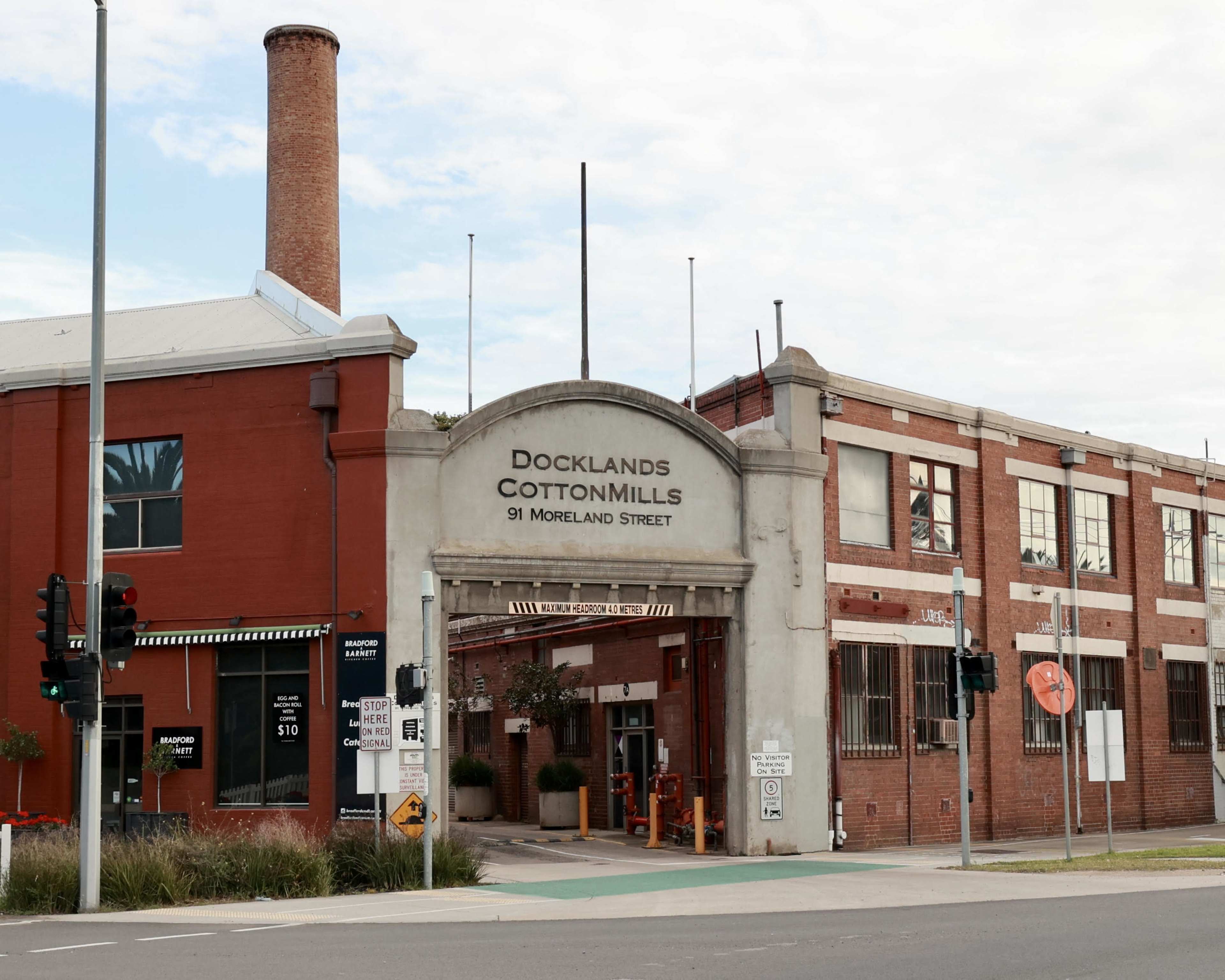 The image shows the entrance to the Docklands Cotton Mills building on Moreland Street, featuring a large archway and a brick structure with a smokestack in the background.