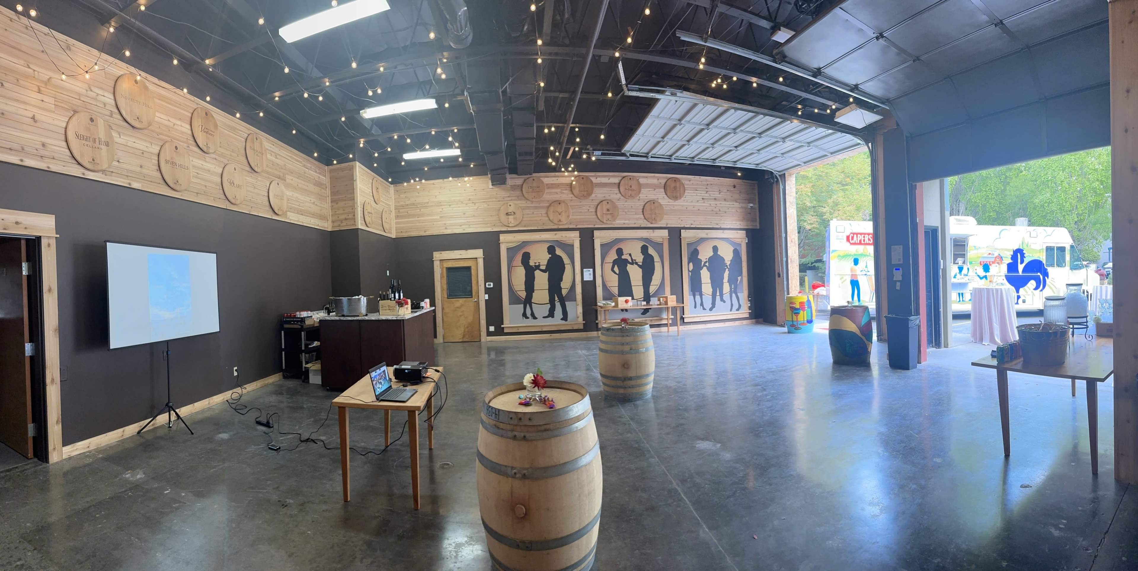 The interior of a rustic event space features wooden barrel tables, a large projection screen, and silhouette artwork on the walls.