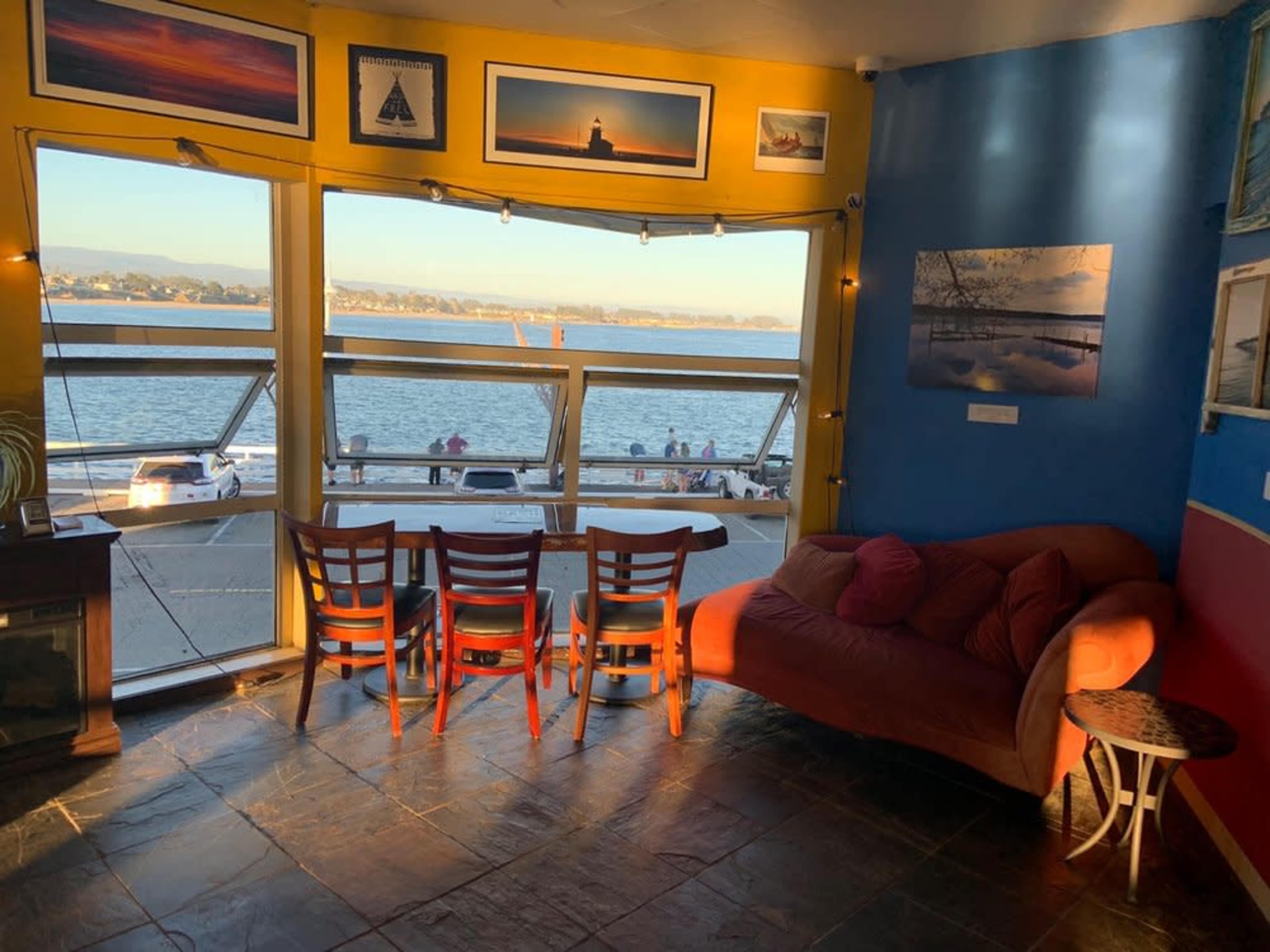 The scene depicts a cozy interior with three chairs and a couch facing large windows that overlook a waterfront view, illuminated by natural light.