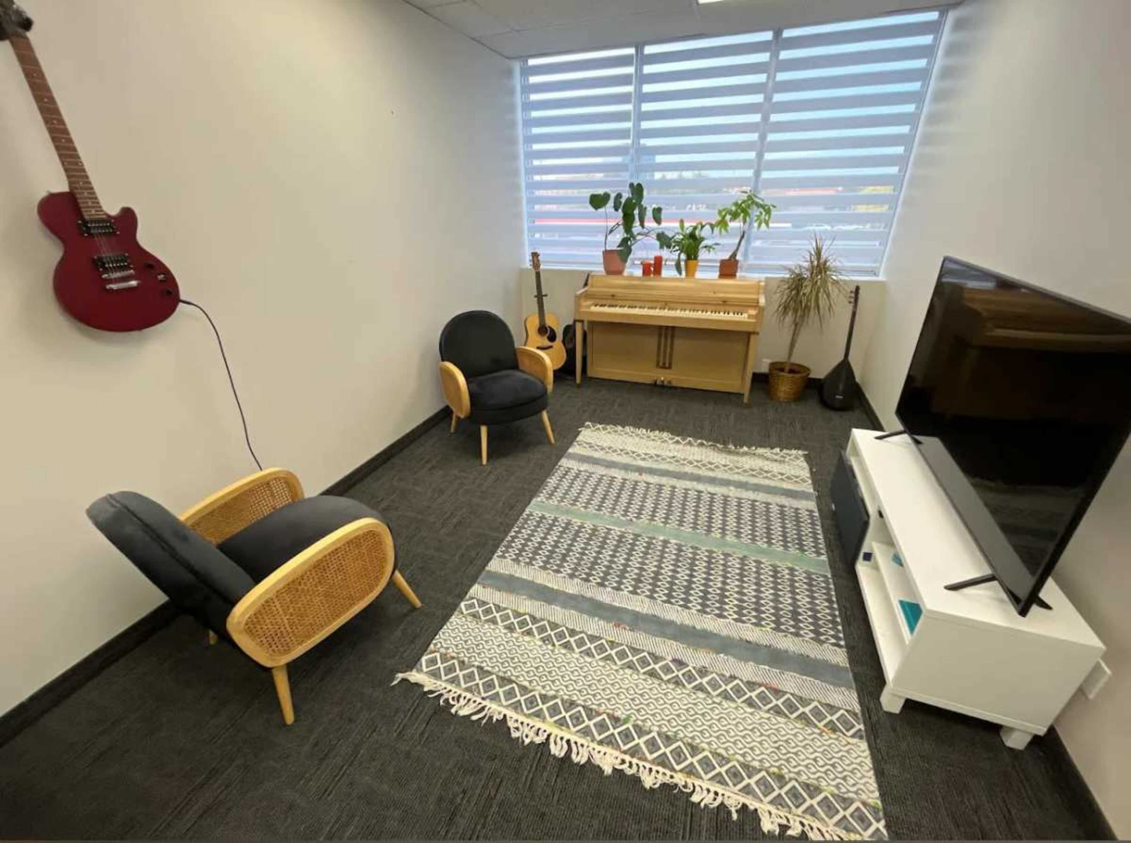 The image shows a small, minimalist room featuring two chairs, a piano, a guitar on the wall, a plant, a rug, and a television.