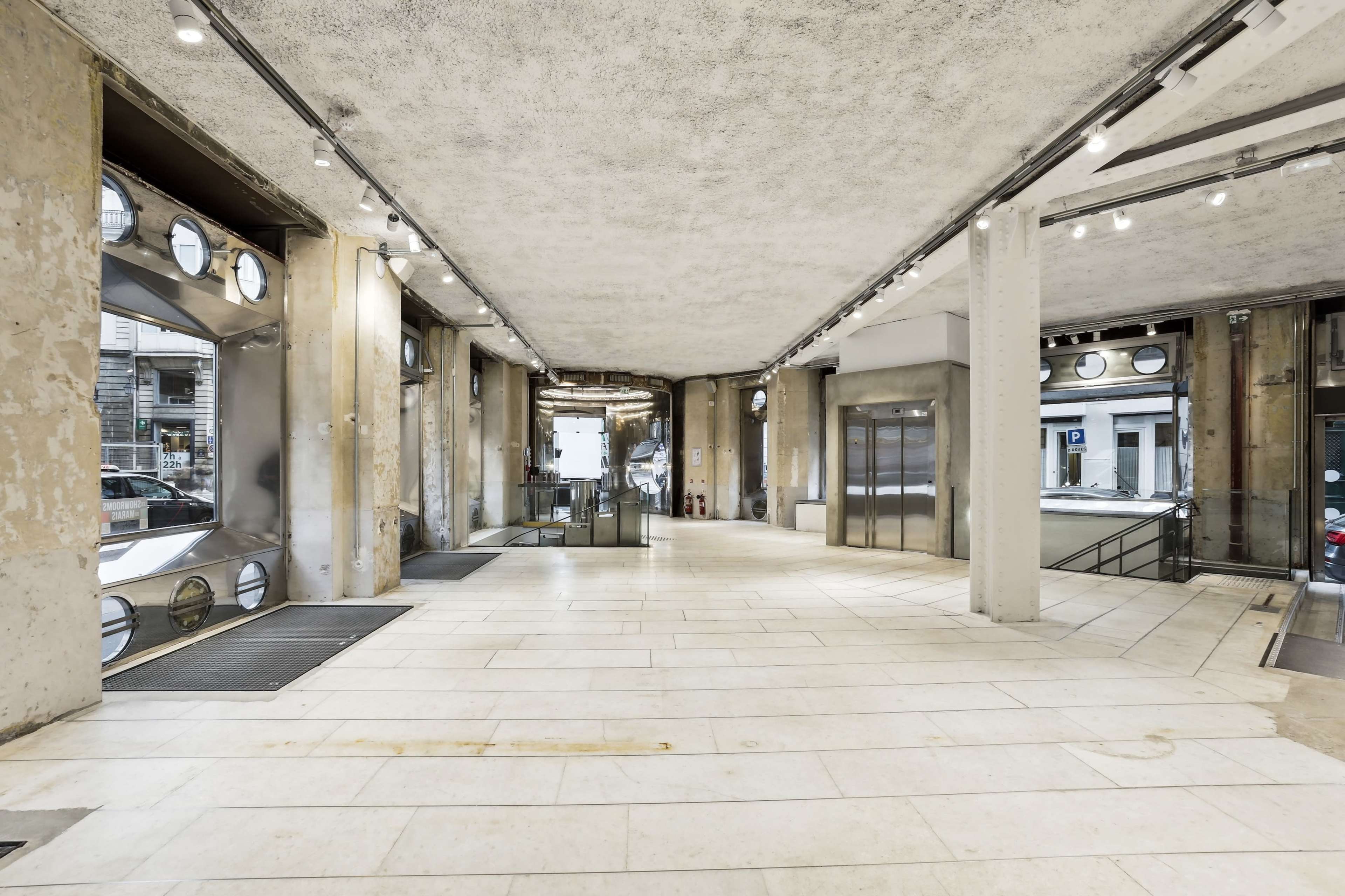 The image shows an industrial-style interior space with exposed concrete walls, large windows, and elevators, illuminated by overhead lighting.