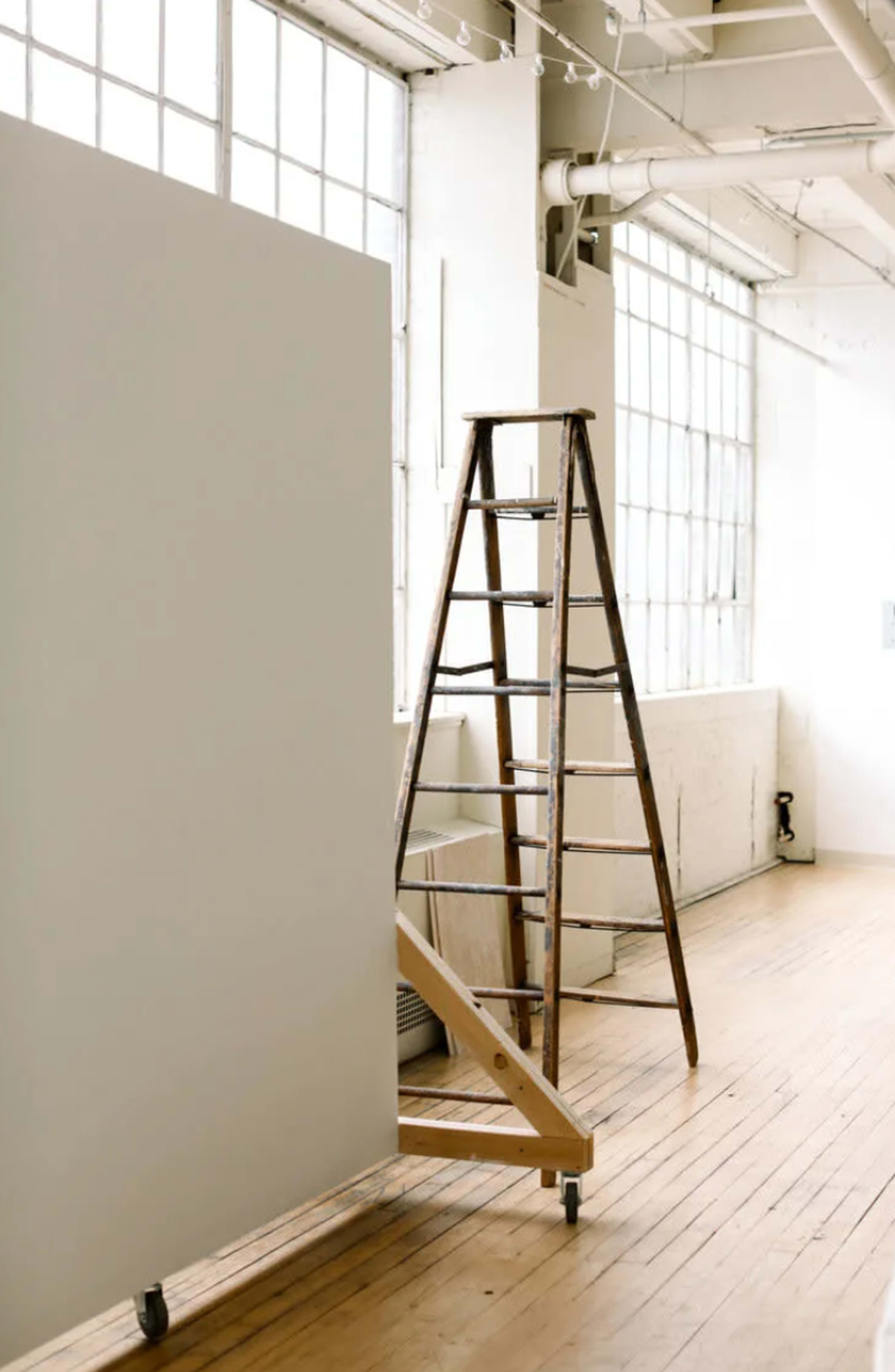 A wooden ladder is positioned next to a large white panel in a well-lit room with large windows and wooden flooring.