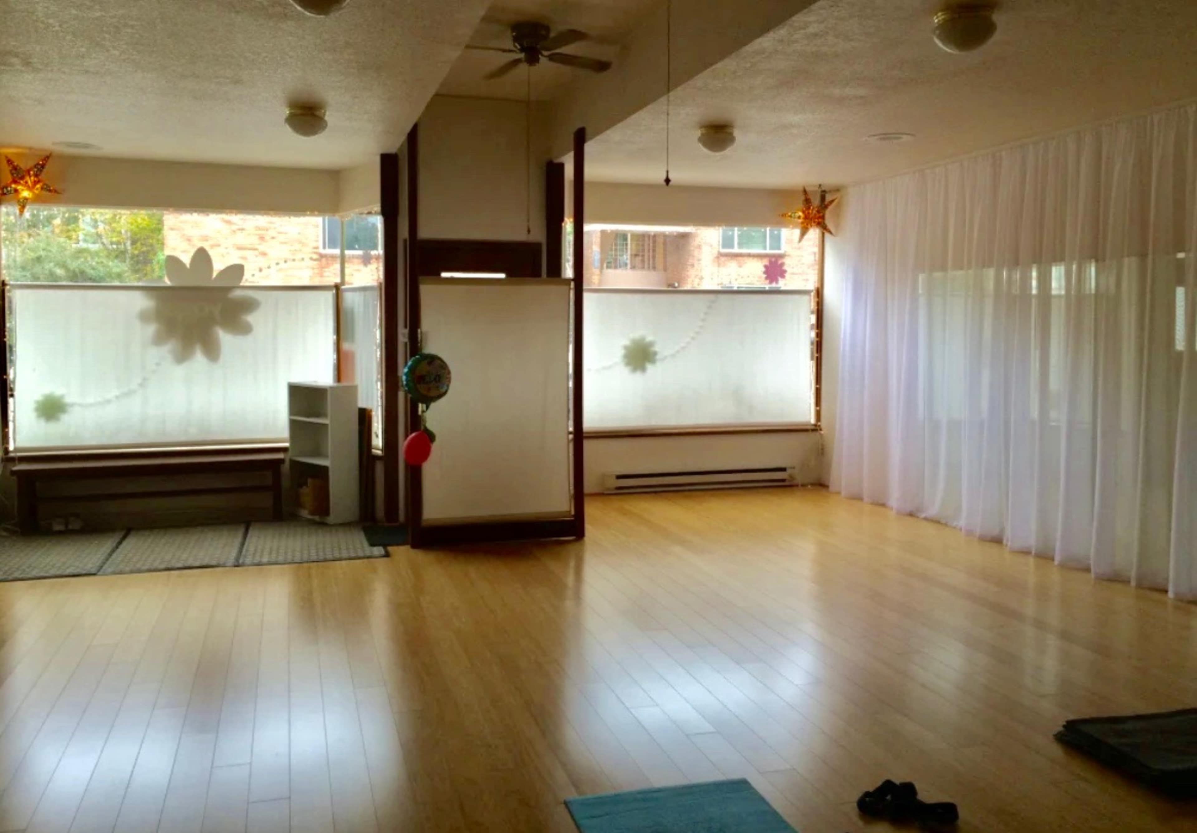 A spacious room with wooden flooring, large windows covered by sheer curtains, and a simple partition separating different areas.