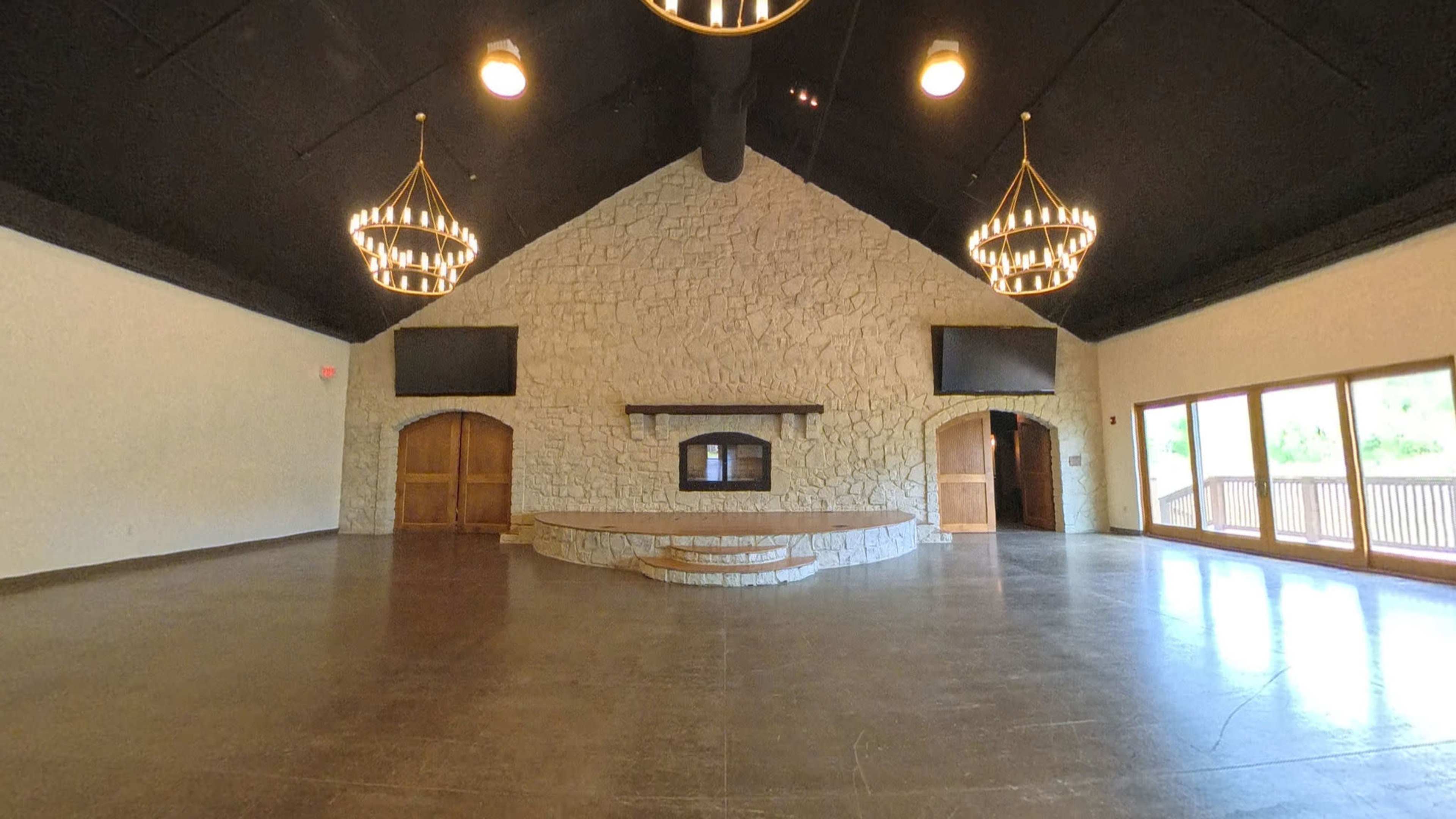 The image shows a spacious indoor venue with a stone fireplace, chandeliers hanging from the ceiling, and large windows leading to an outdoor area.
