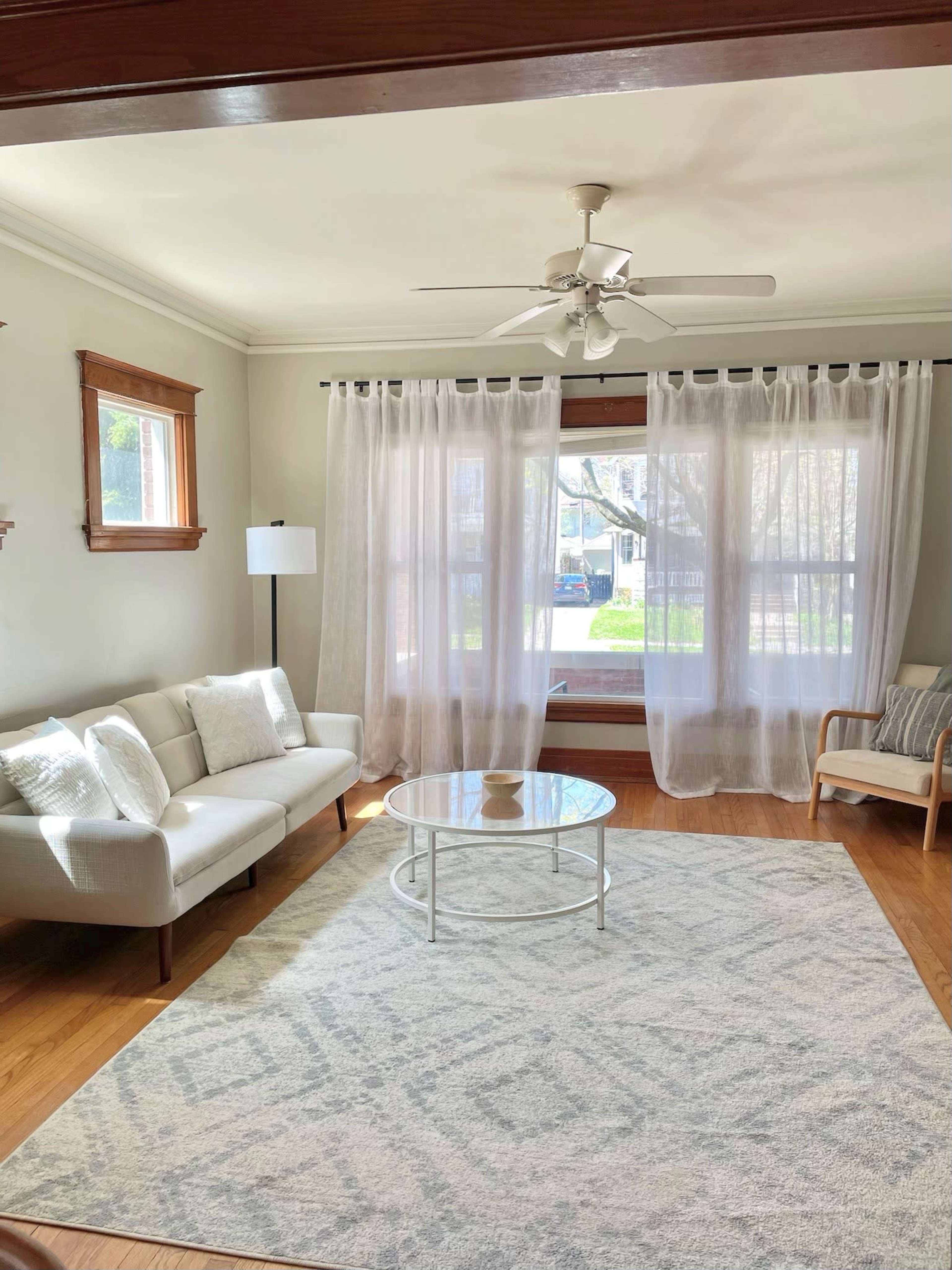 A bright living room features a white sofa, a round glass coffee table, a patterned rug, and large windows with sheer curtains.
