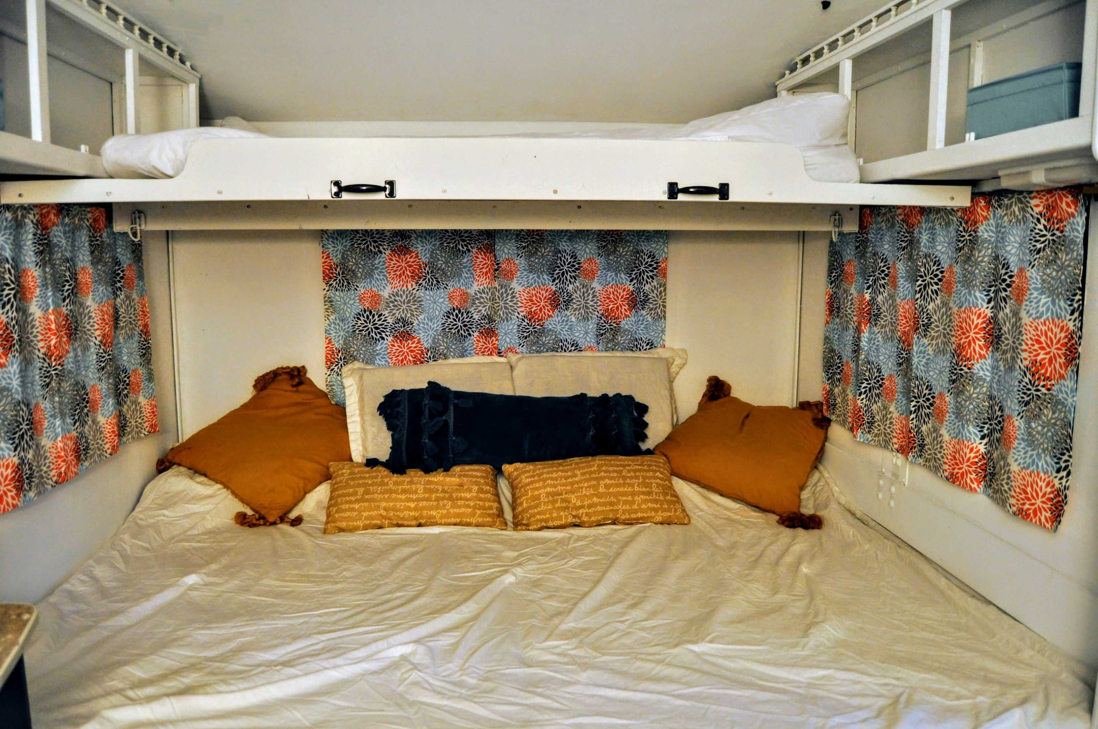 The image shows a cozy sleeping area with a lower bed covered in white bedding and adorned with various decorative pillows, flanked by curtains with a floral pattern.