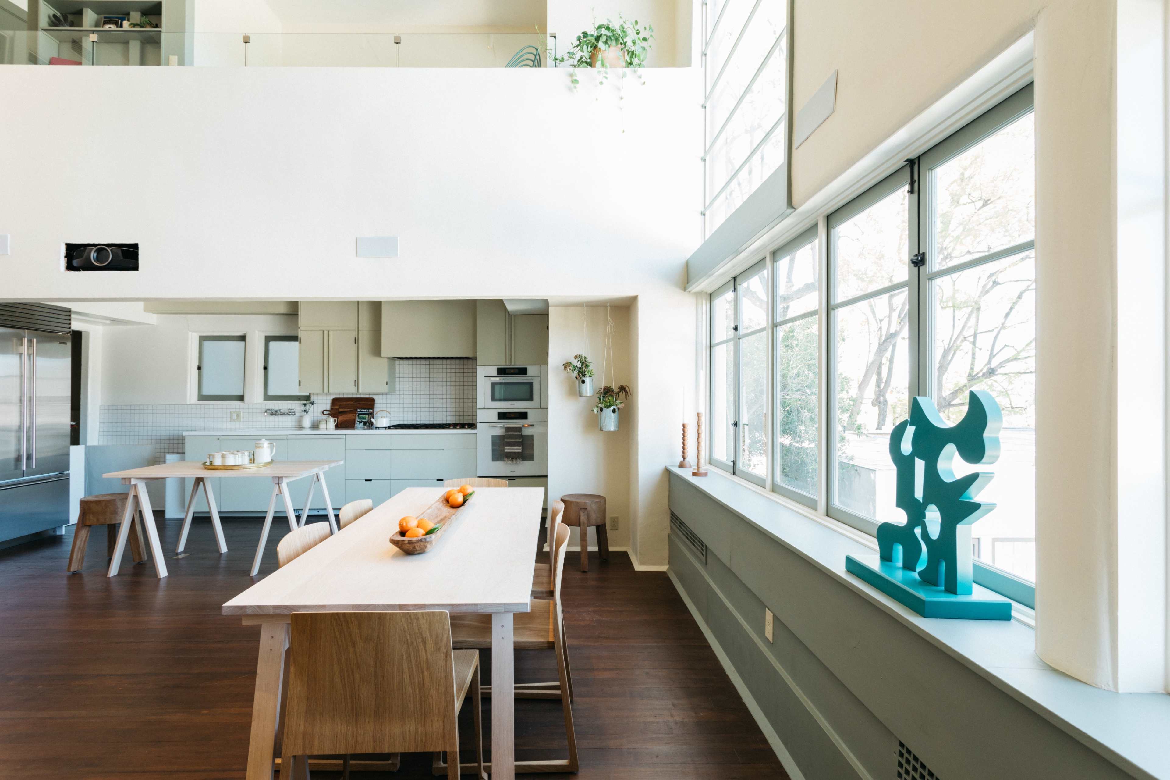 A bright kitchen and dining area features a wooden table with chairs, a countertop with appliances, and large windows letting in natural light.
