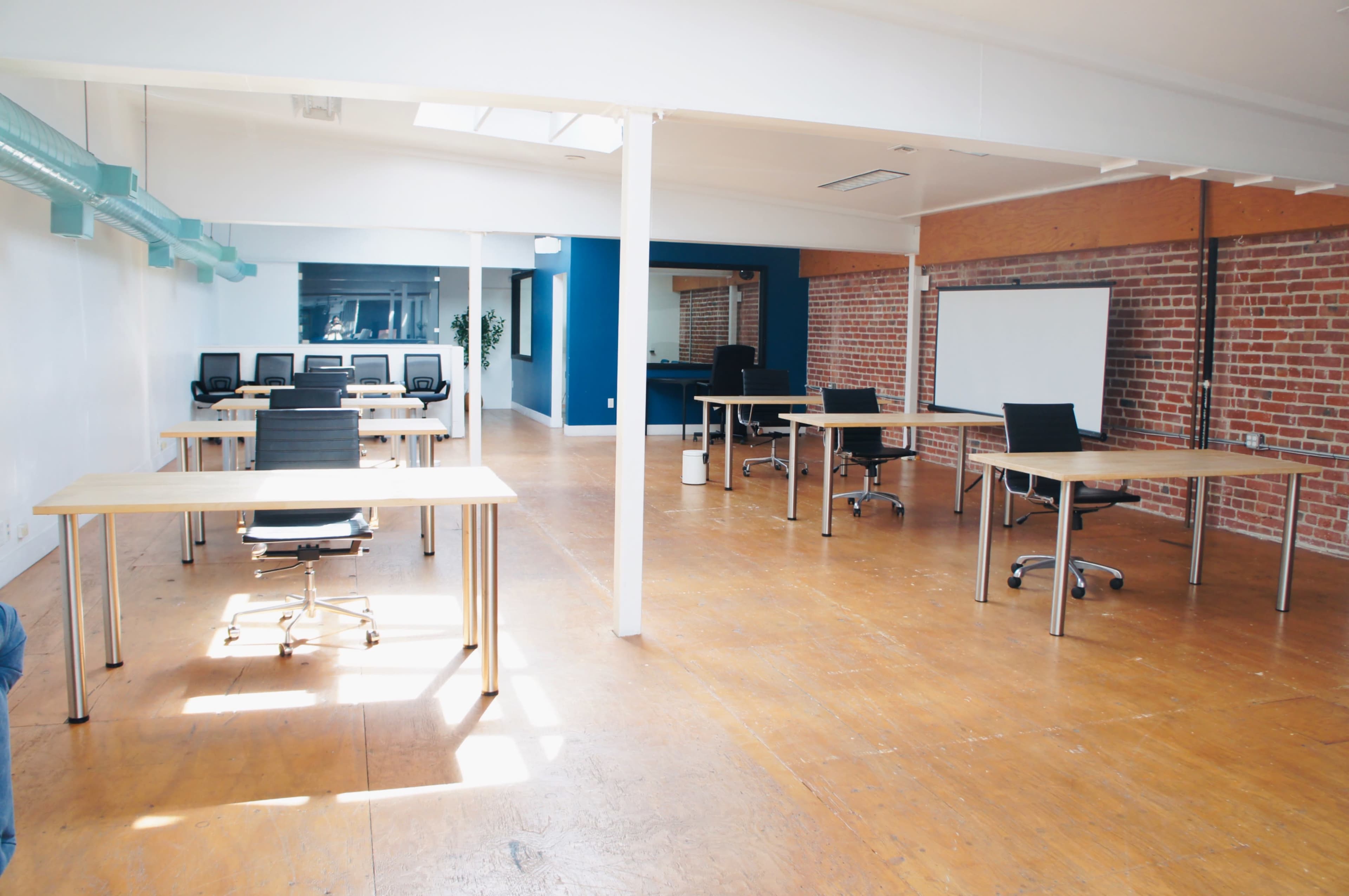 The image shows a spacious office setting with several wooden desks and chairs arranged, along with a presentation screen and large windows allowing natural light.