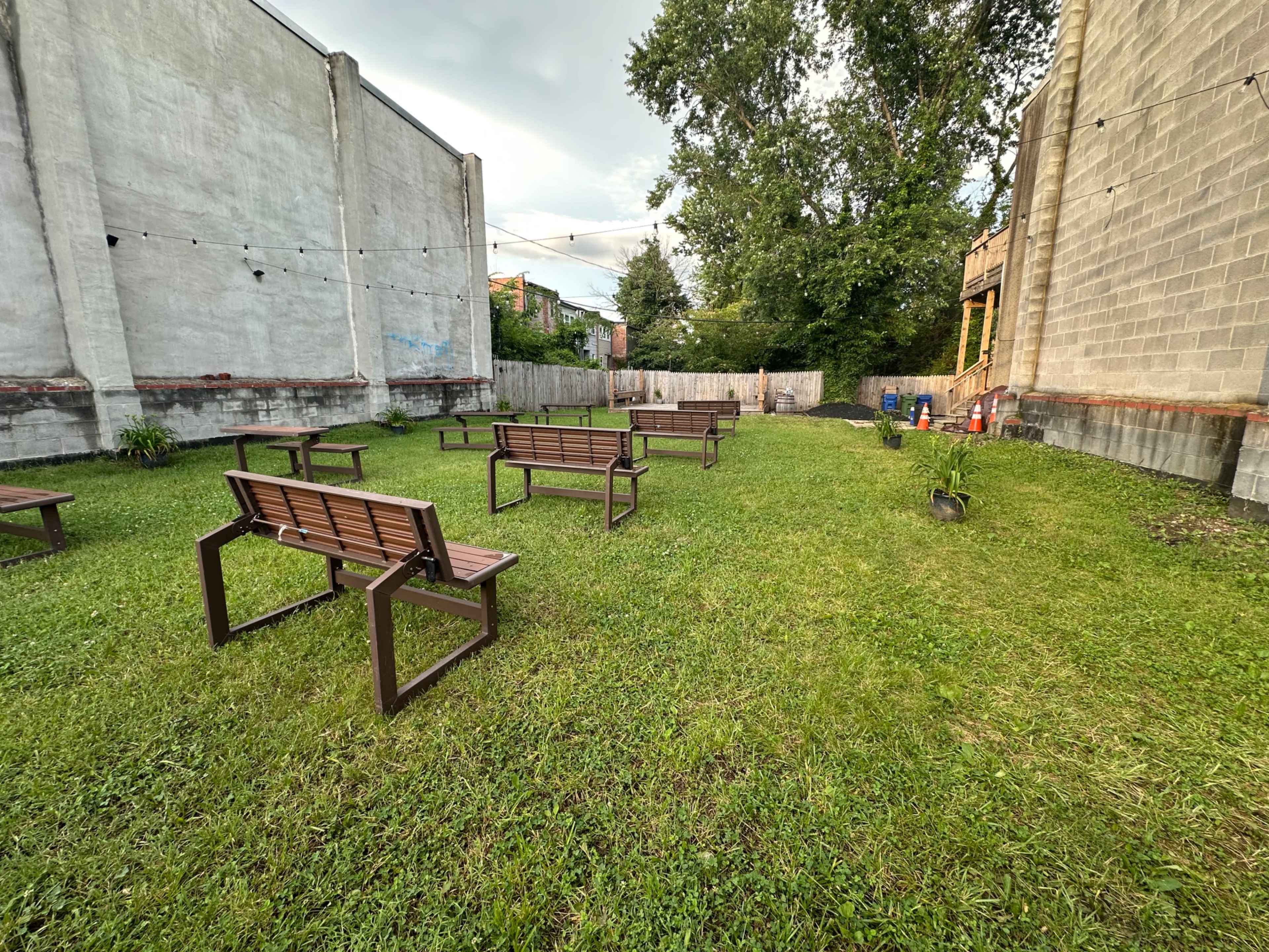 The image shows a grassy outdoor area with several wooden benches arranged in a grid pattern, and surrounding walls of buildings on two sides.