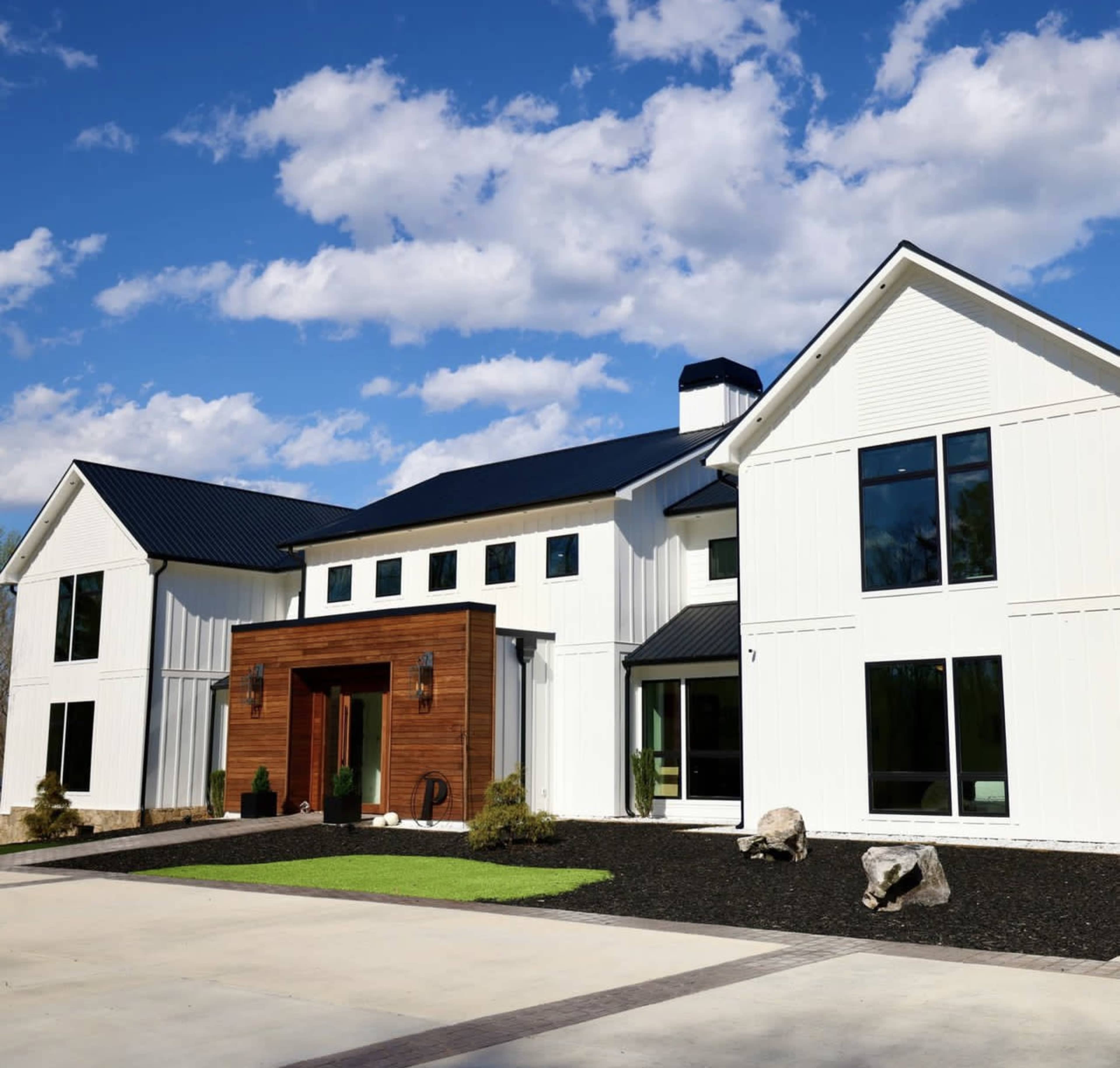 A modern two-story house with a white exterior, a wooden front entrance, large windows, and landscaped grounds featuring rocks and grass.