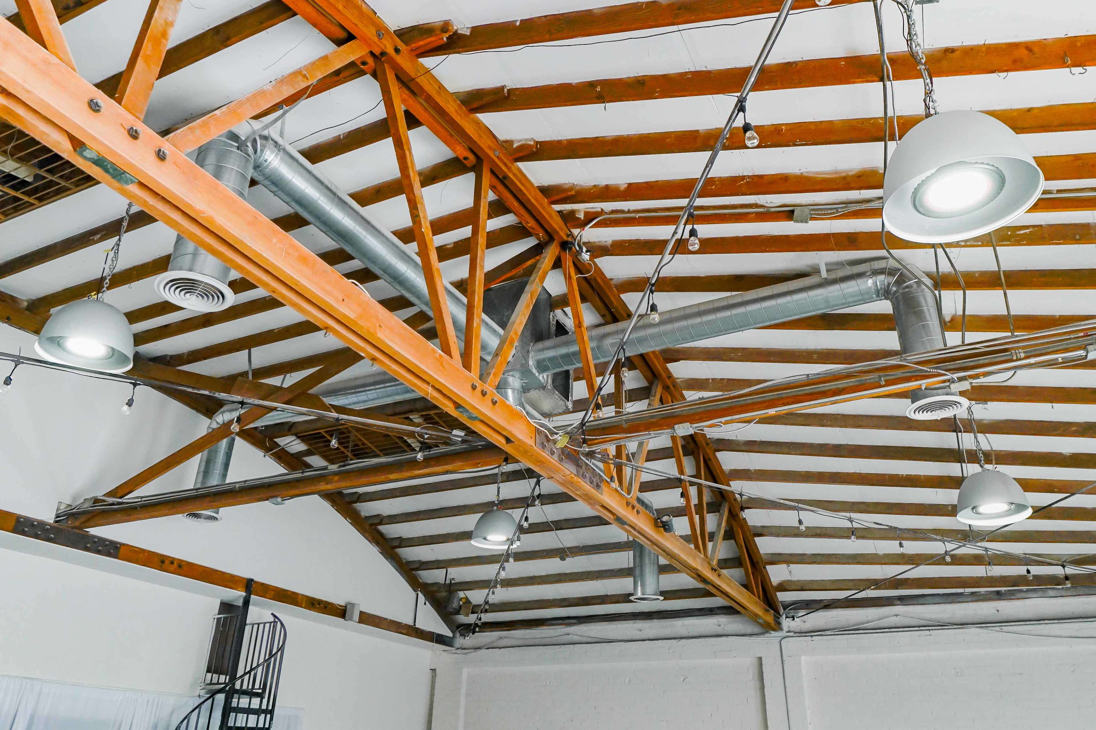 The image shows a loft space with exposed wooden beams and metal ductwork, featuring pendant lights hanging from the ceiling.