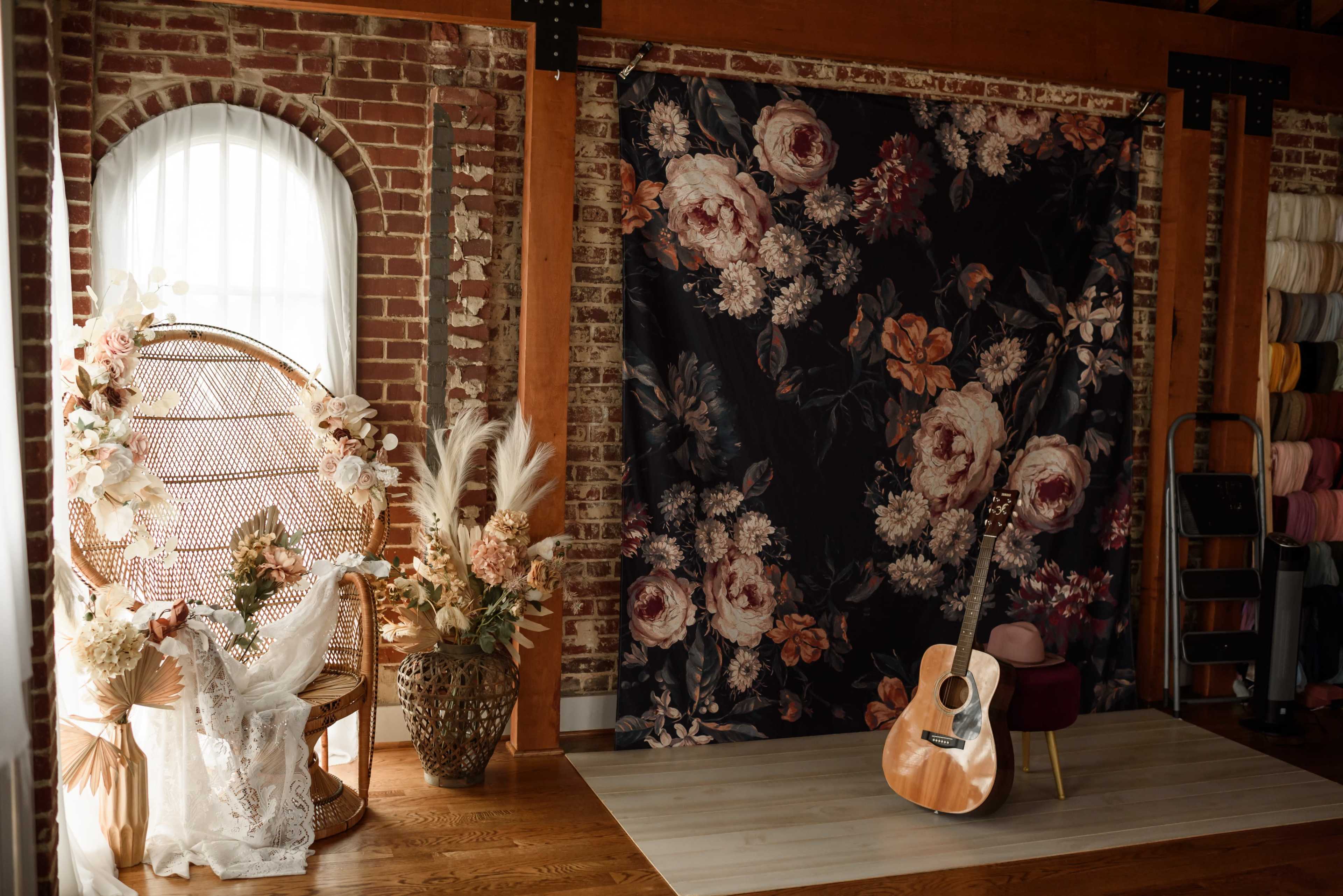 The image features a cozy indoor setting with a wicker chair adorned with flowers, a floral backdrop, a potted plant, and an acoustic guitar leaning against a chair.