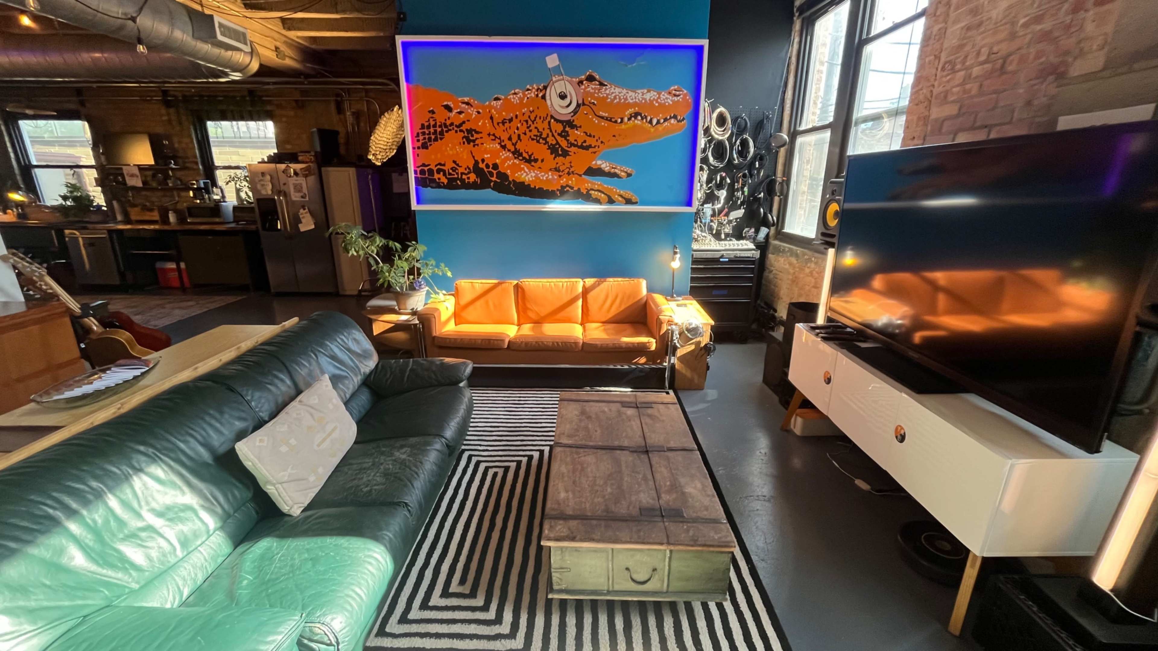 The image shows a stylish urban living room with a green leather couch, an orange sofa, a wooden coffee table, and a large artwork of an alligator on the wall.