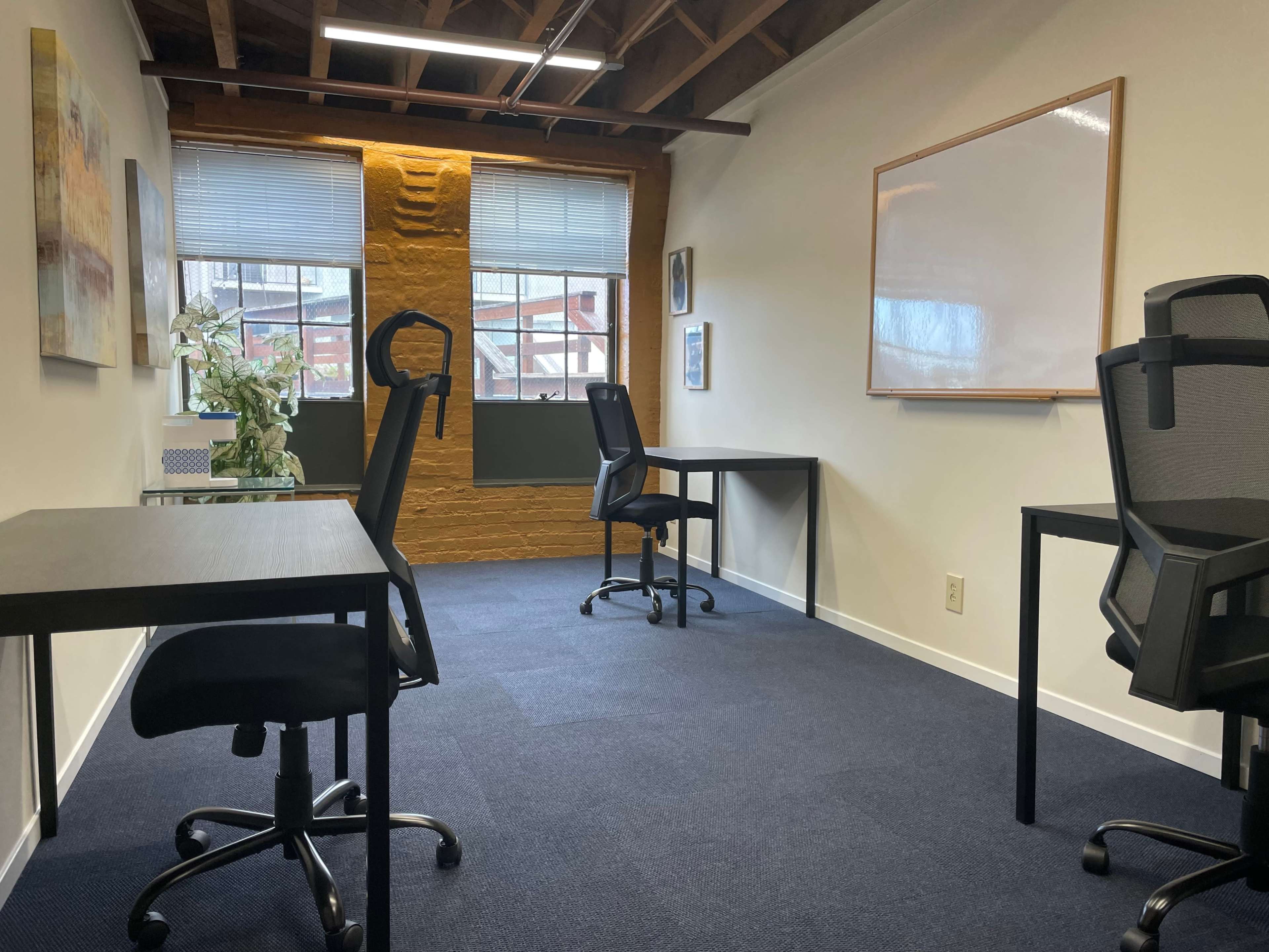 The image shows a partially furnished office space with two desks and chairs, situated in a room with wooden beams and large windows.