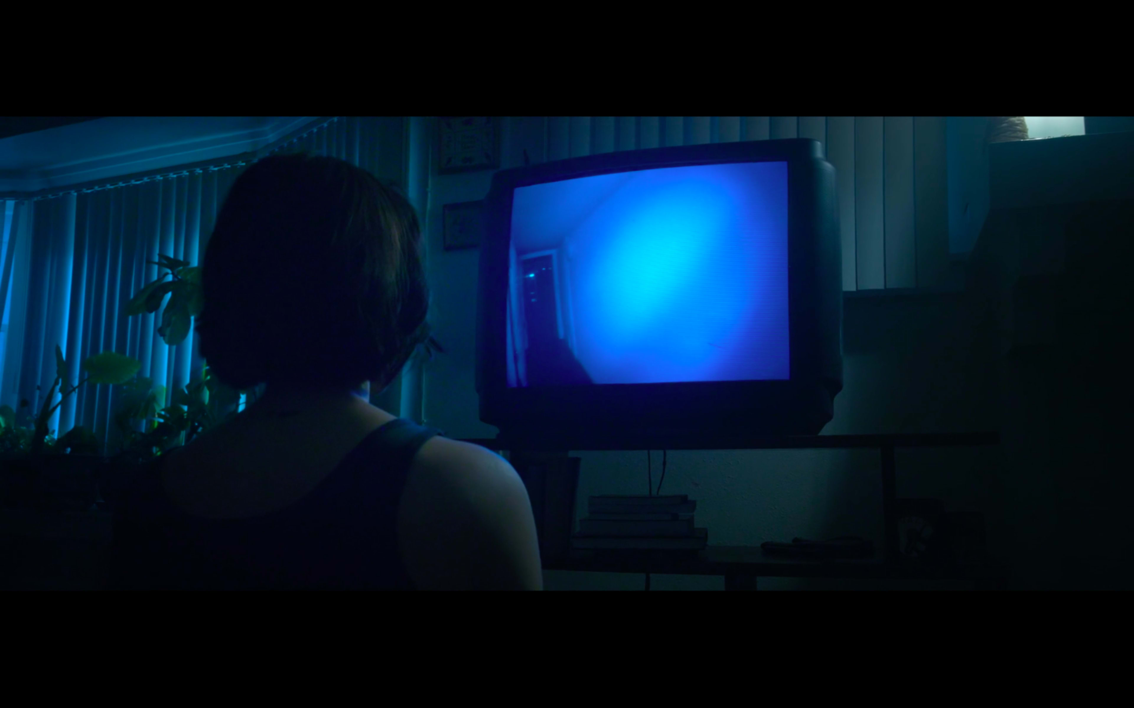 A person sits in front of a vintage television displaying a bright blue image, with the dimly lit room visible behind them.