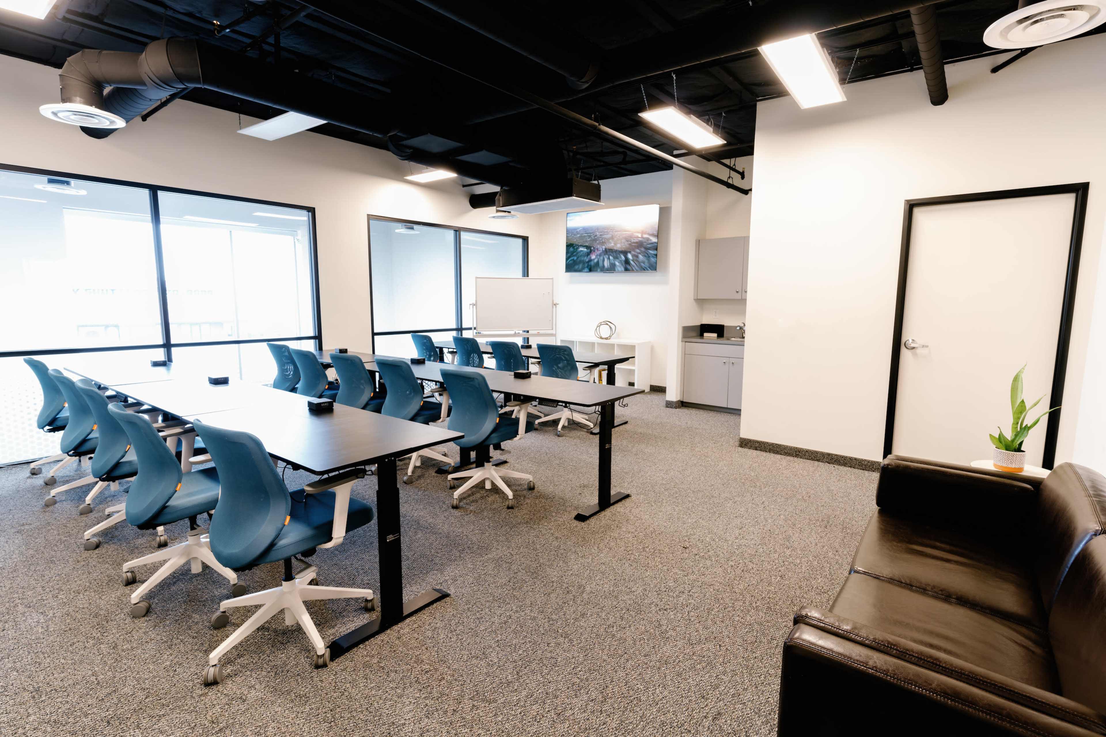 A modern conference room features a long table with blue chairs, large windows providing natural light, and a sofa in one corner.