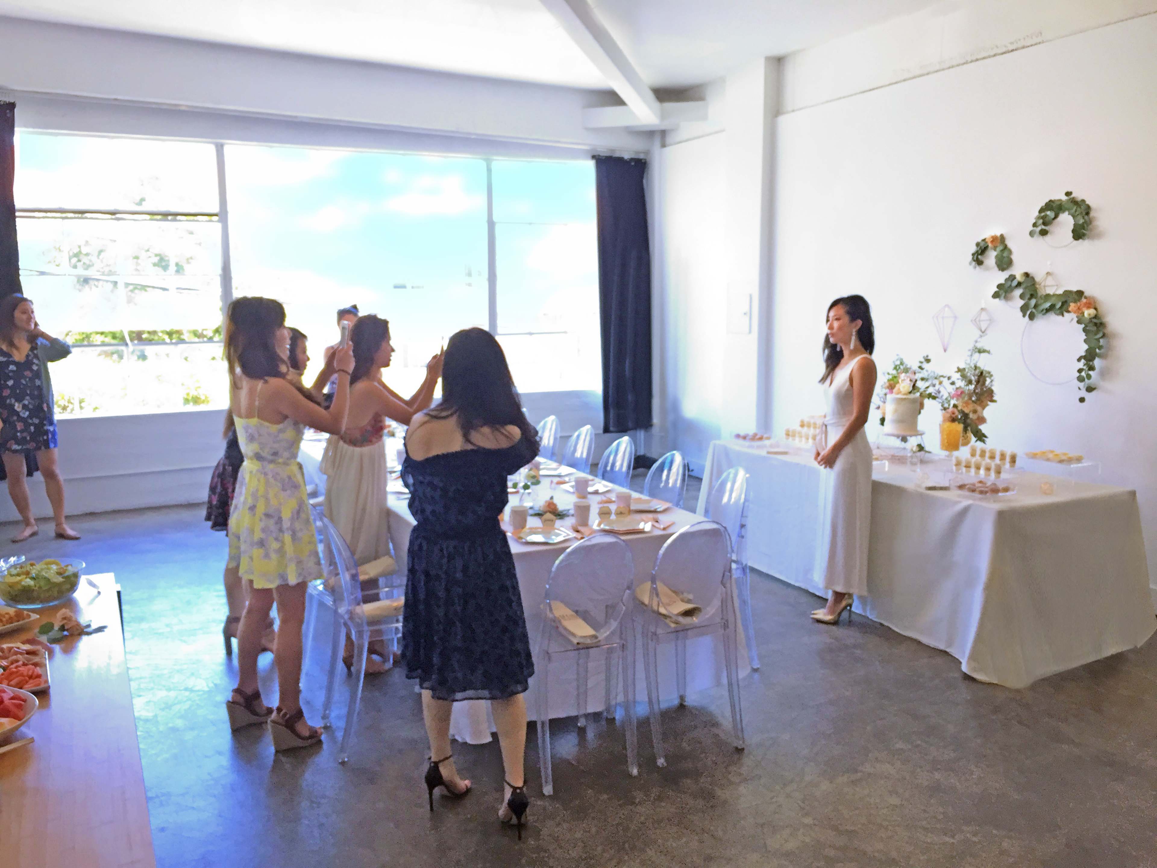 A group of women takes photos of a woman standing by a decorated table in a brightly lit room.