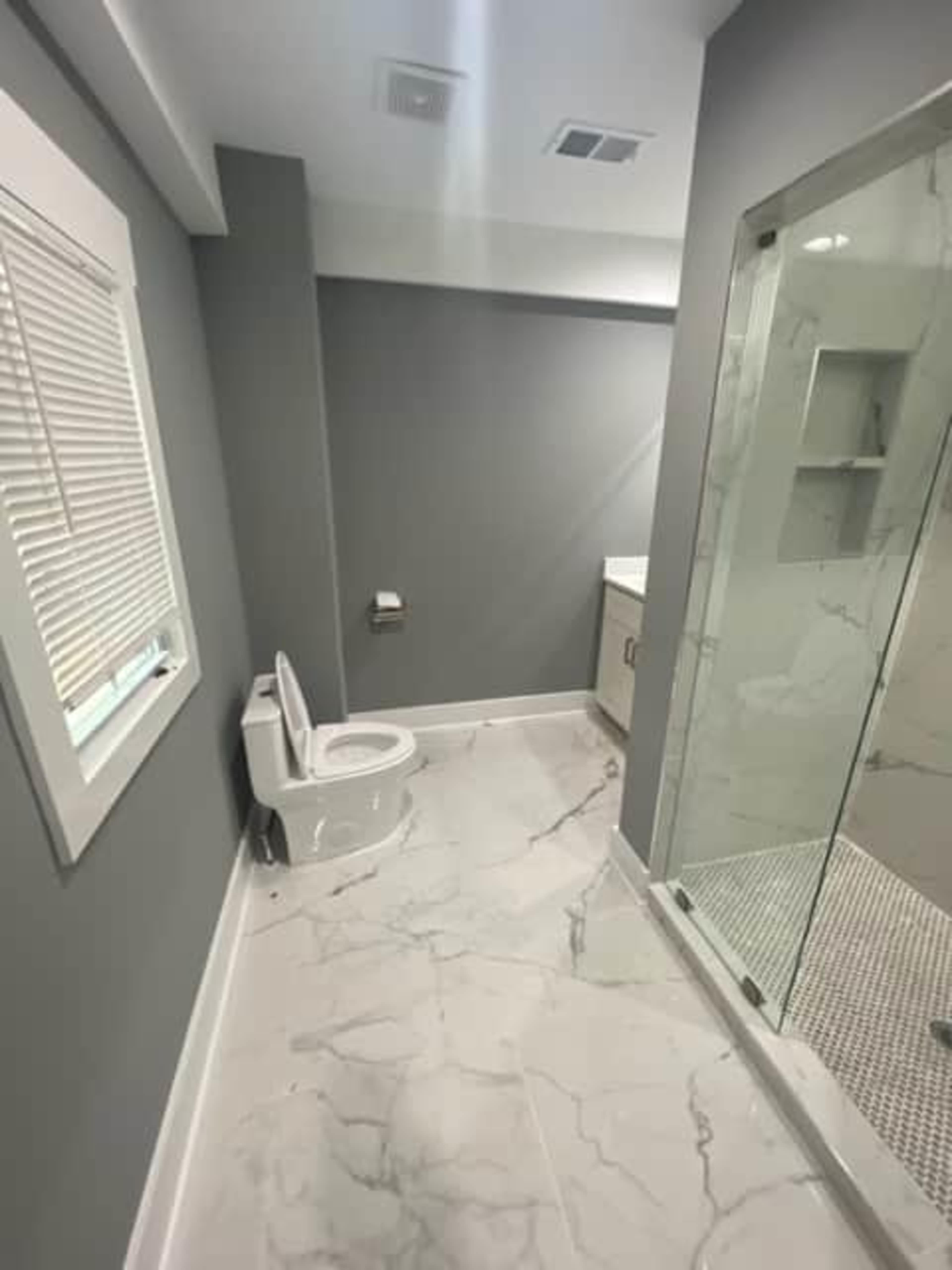 The image shows a modern bathroom featuring a toilet, a shower with a glass door, and a vanity with a countertop, all set against gray walls and marble flooring.
