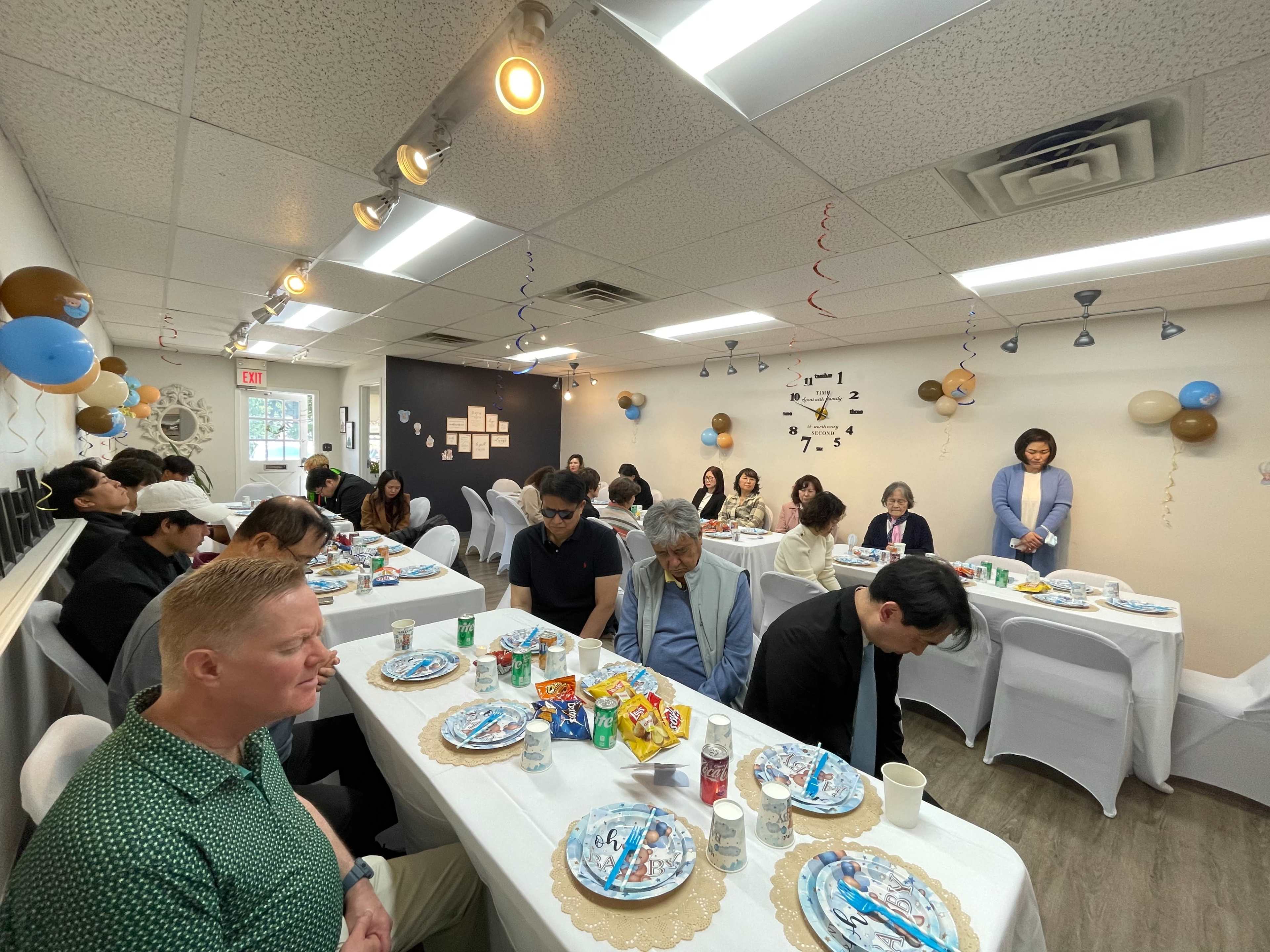 A group of people seated at tables in a decorated room, participating in a gathering with various snacks and drinks on the tables.