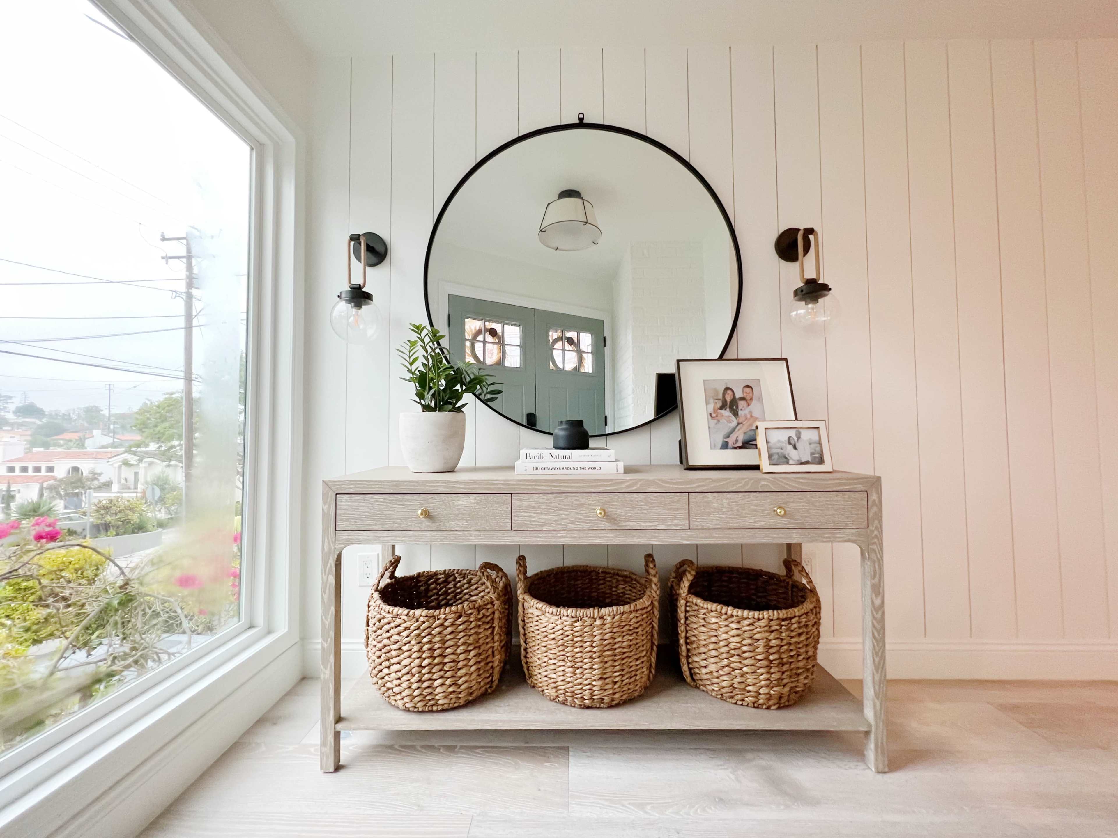 A light-colored wooden console table with three woven baskets underneath and framed photos on top, positioned near a large window with a round mirror above it.