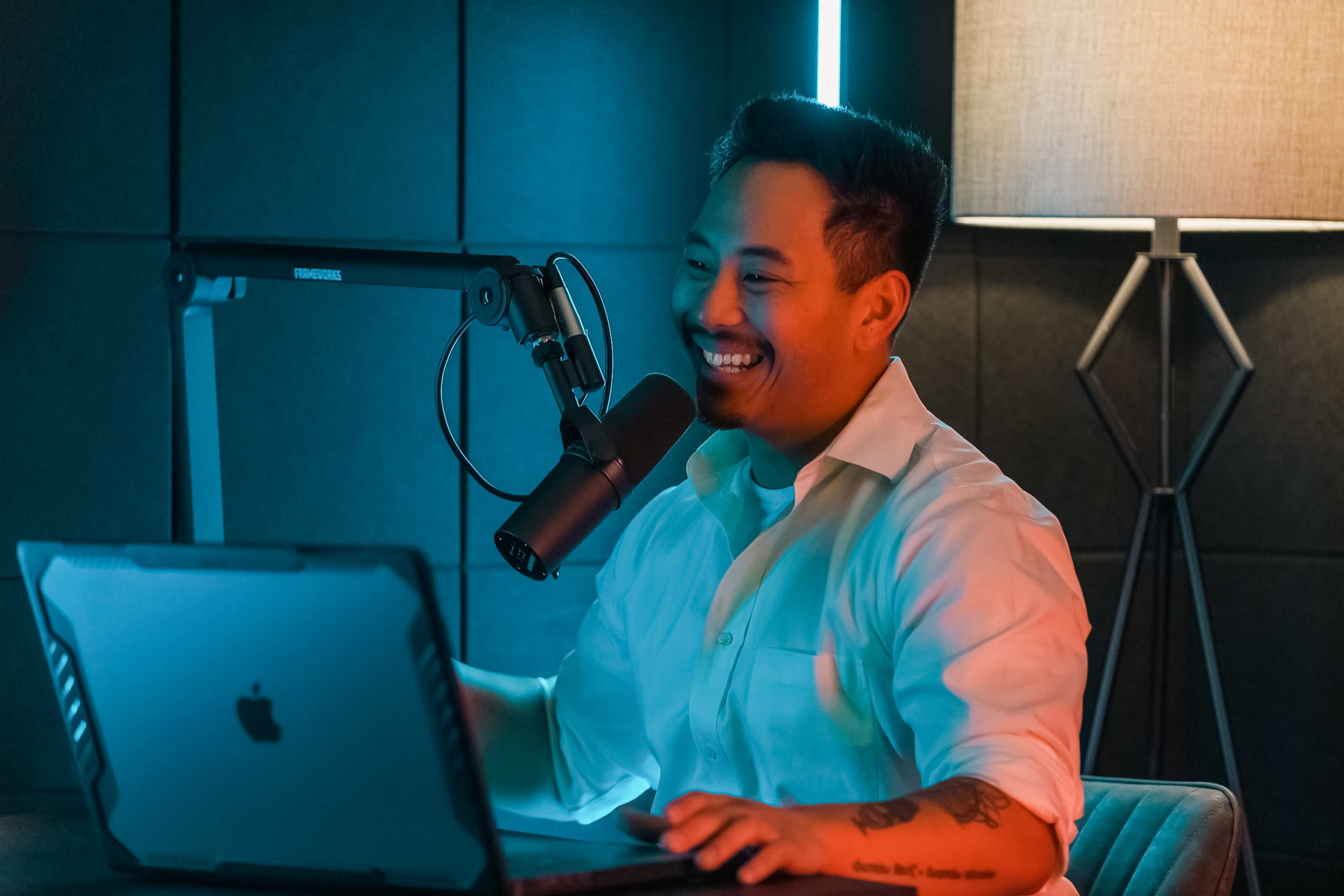 A man with a microphone smiles while sitting at a laptop in a dimly lit room with a lamp.