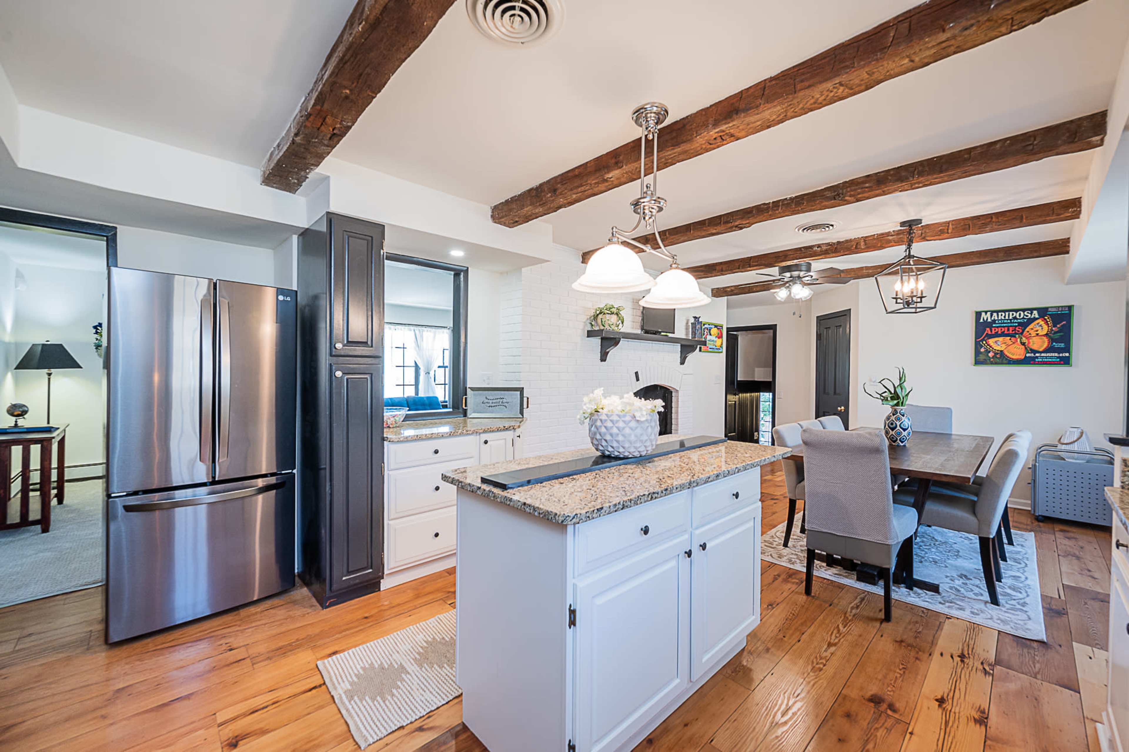 A modern kitchen features a central island with granite countertops, wooden beams on the ceiling, and a dining area with a table and chairs.