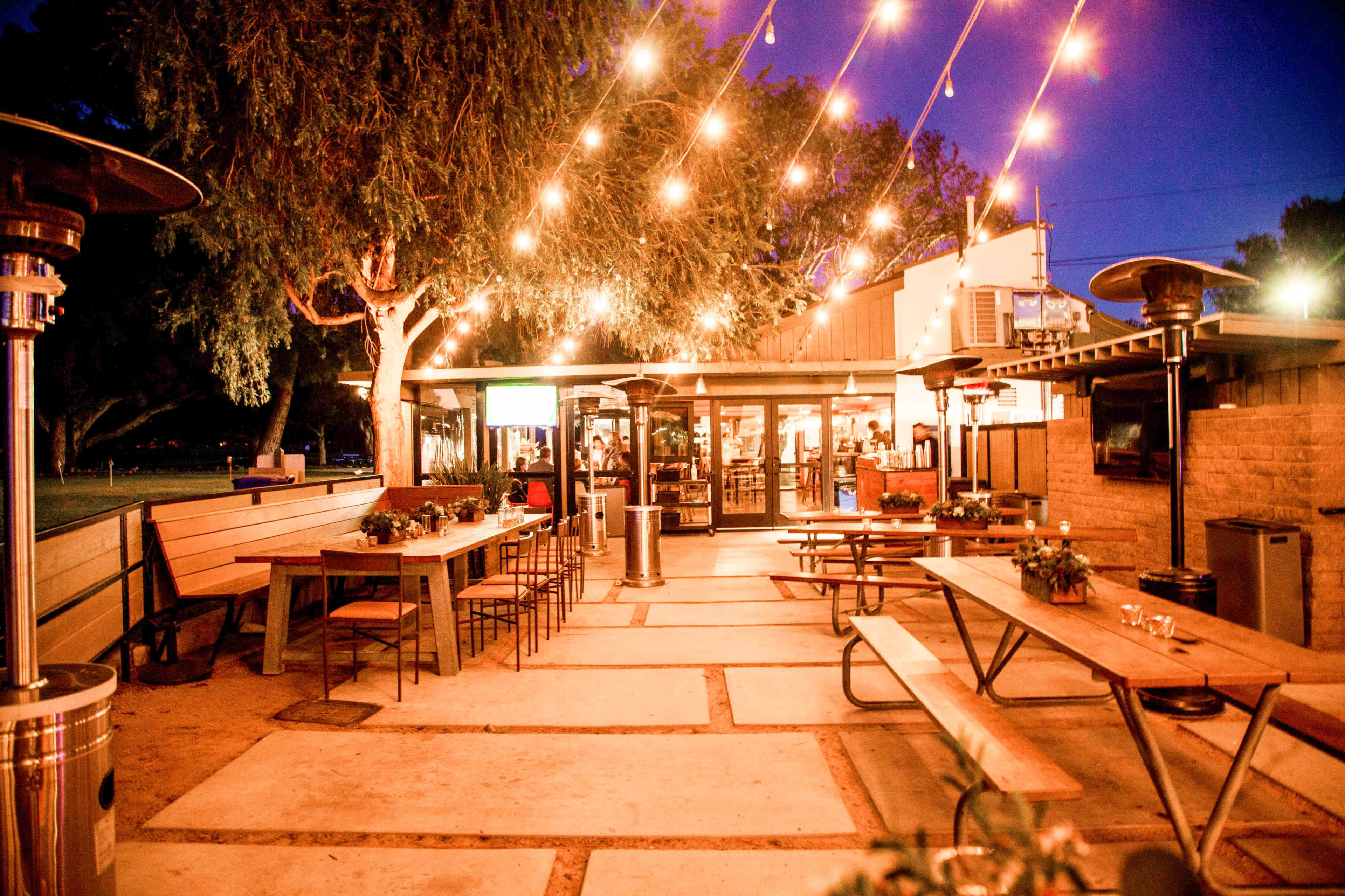 The image shows a lively outdoor dining area with string lights overhead, featuring long communal tables and seating arrangements, alongside a bustling restaurant entrance in the background.