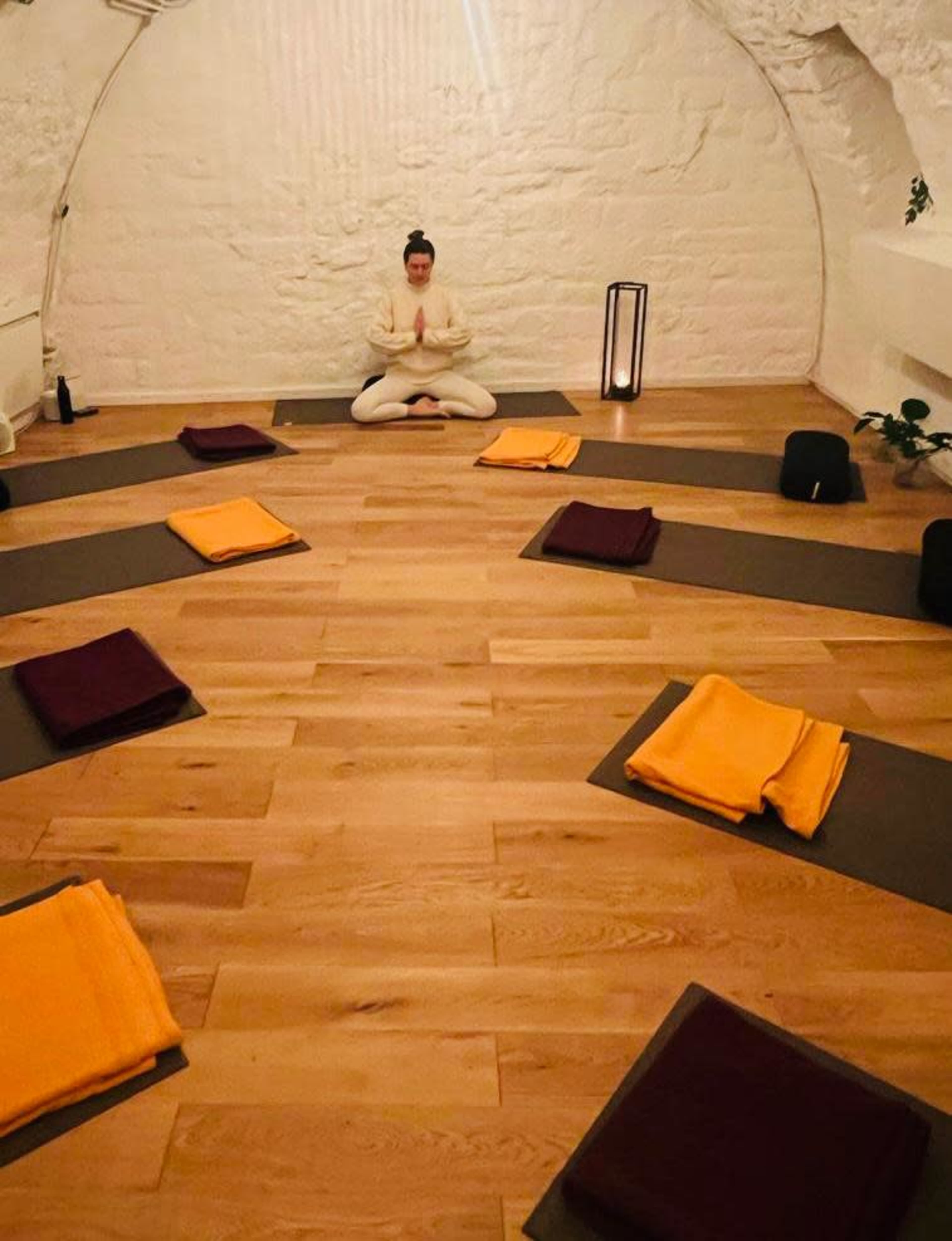 A person sits in a meditative pose surrounded by neatly arranged yoga mats and blankets in a serene, softly lit room.