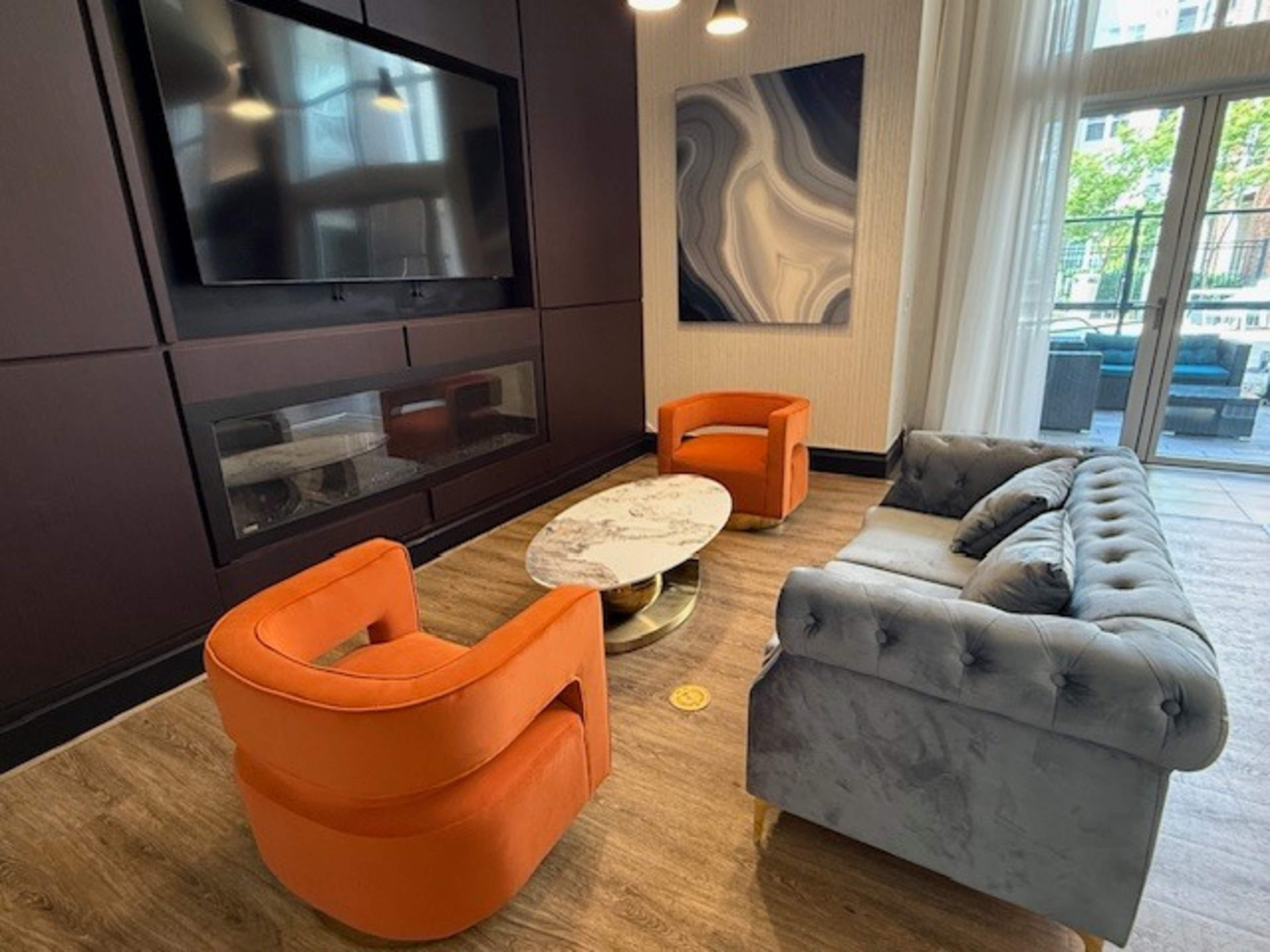 The image shows a modern lounge area featuring a gray tufted sofa, two orange armchairs, a marble coffee table, and a large wall-mounted television.