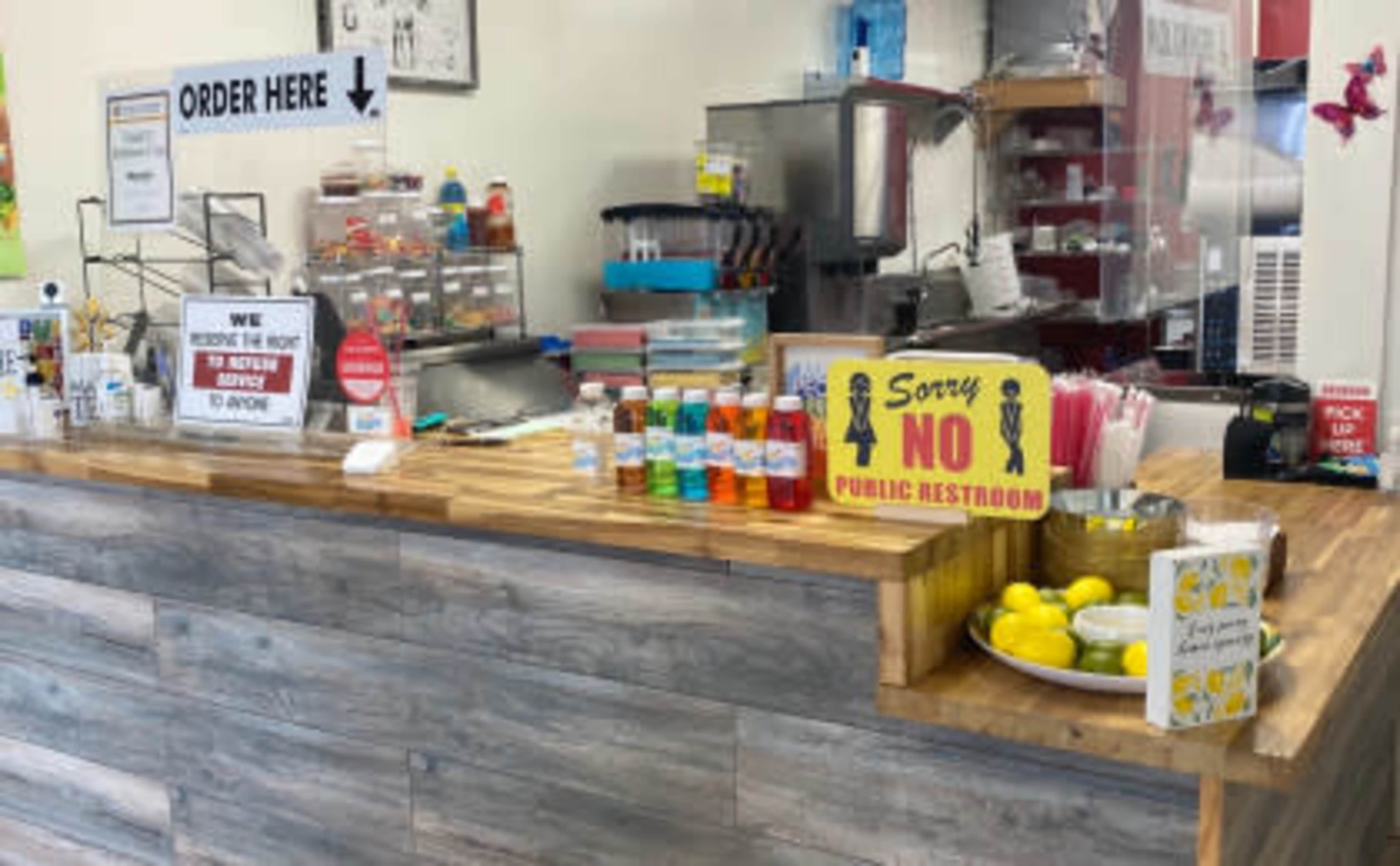A food counter with a wooden surface, bottles of drinks, a sign indicating to order here, and a notice about public restroom restrictions.