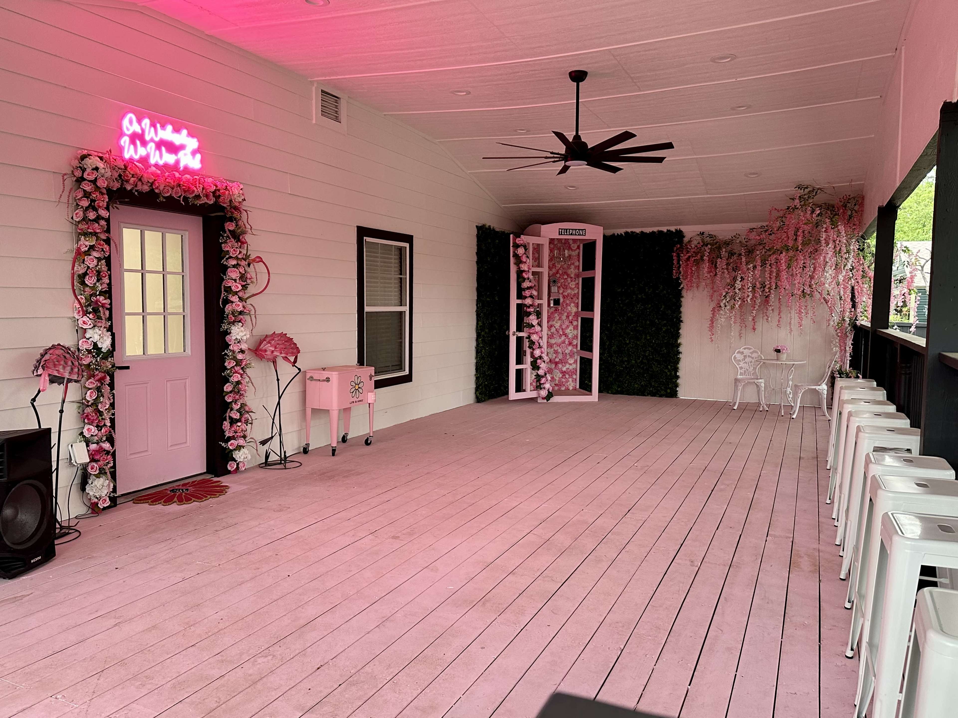 A decorated outdoor porch with pink walls, floral arrangements, seating, and a vintage phone booth.