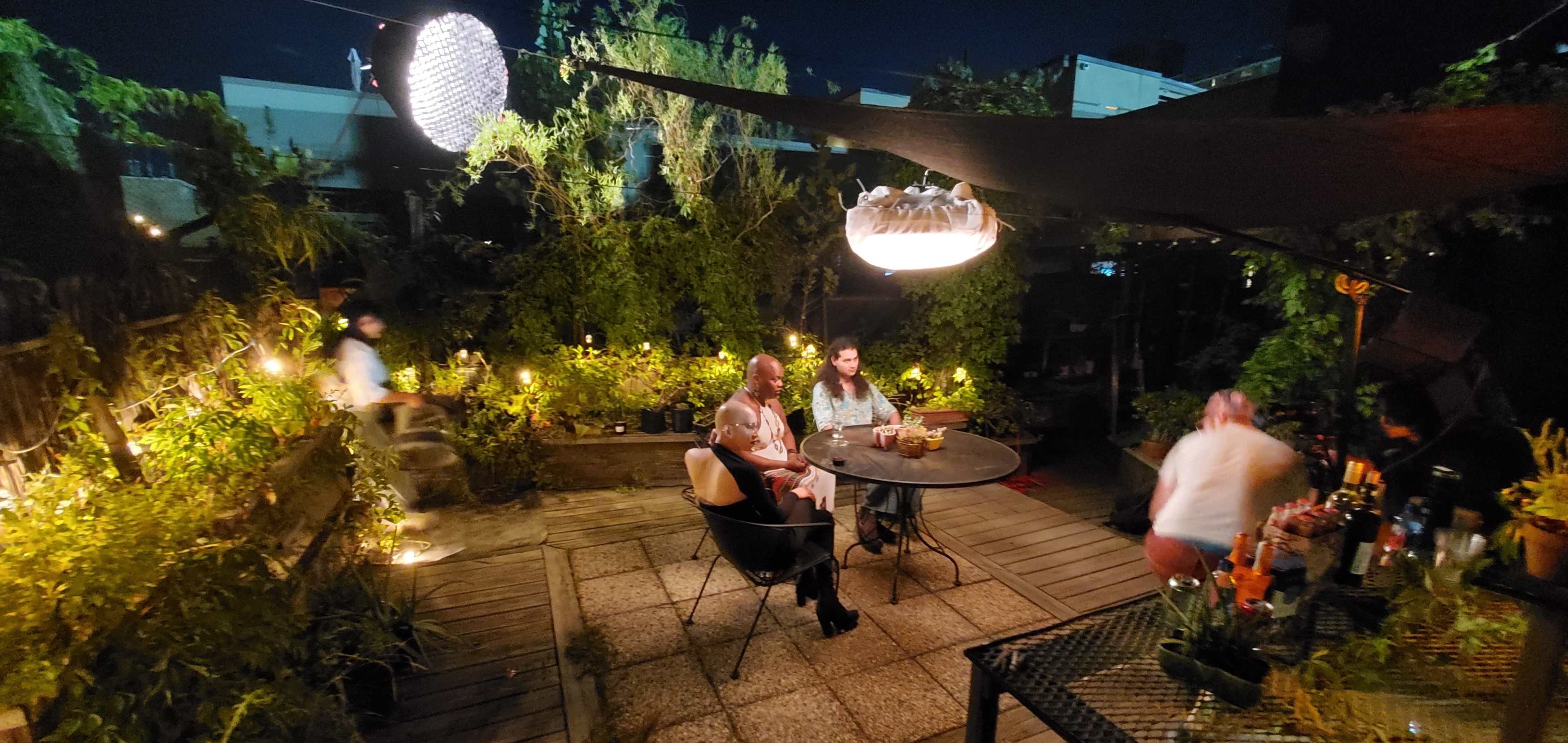 A night scene in a rooftop garden with several people sitting around a table surrounded by plants and soft lighting.