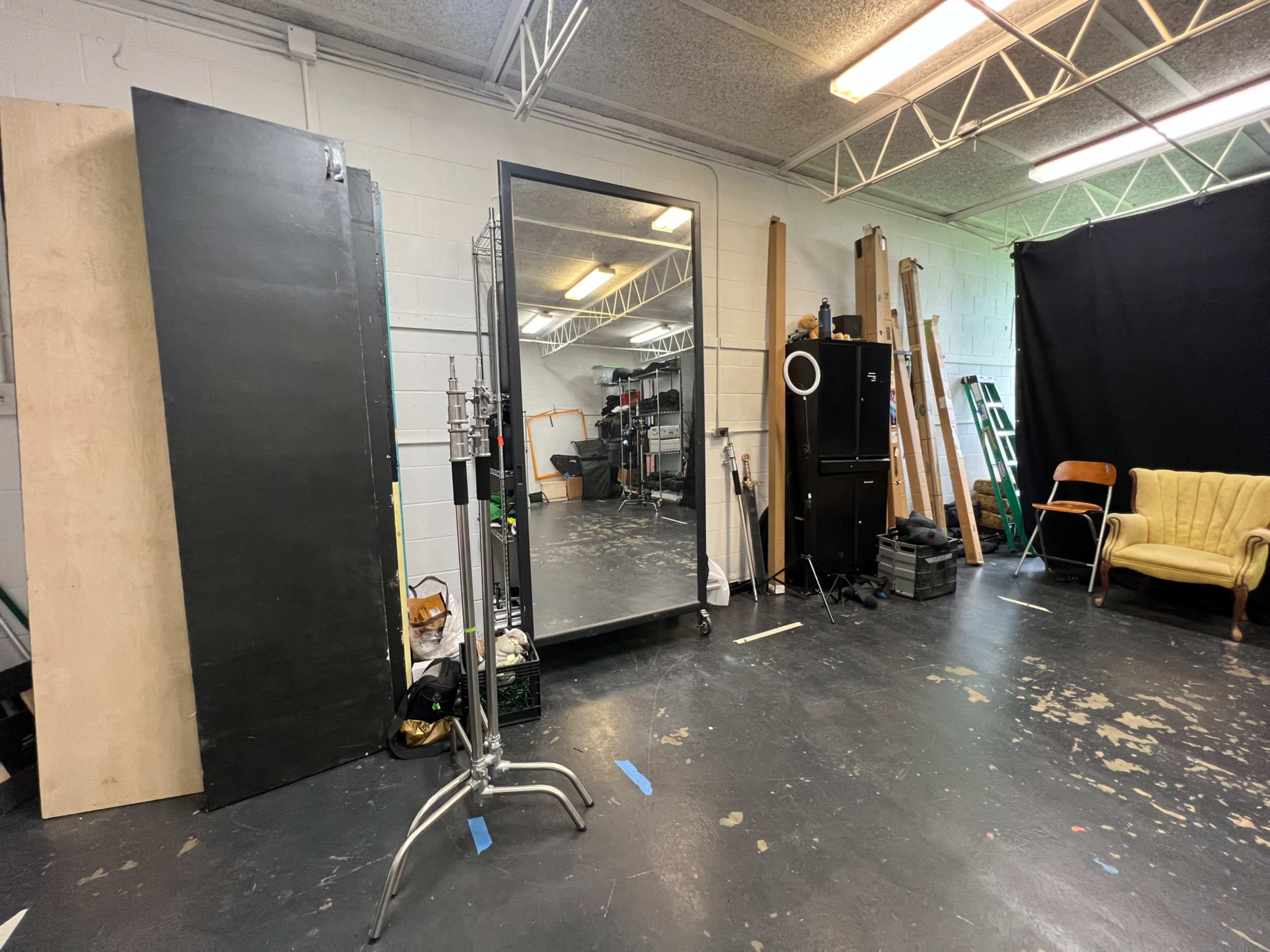 The image shows a studio space with a large mirror, a yellow chair, various pieces of wood, equipment, and a black backdrop.