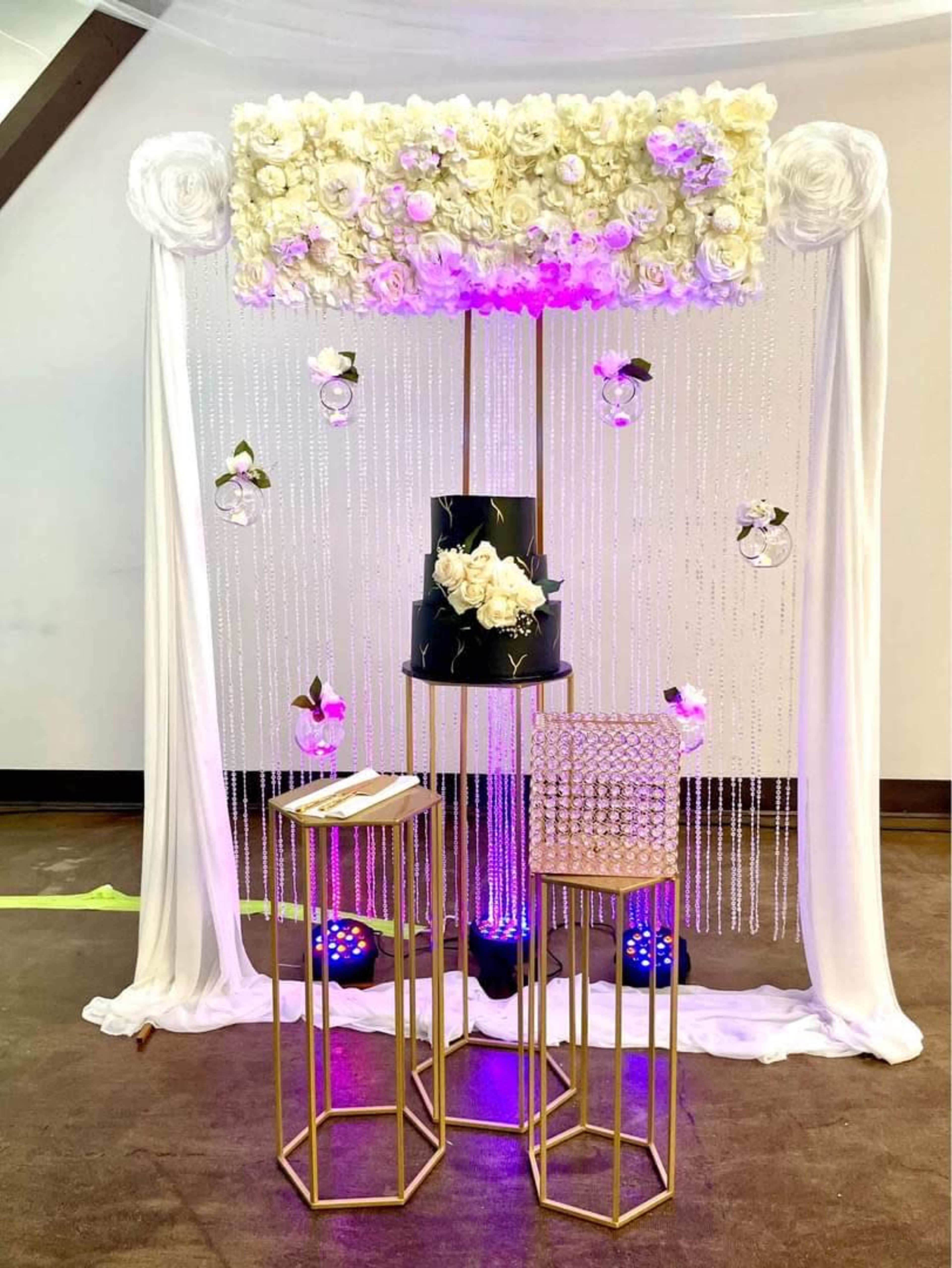 A decorated wedding cake display with a floral backdrop, draped fabric, and hanging glass decorations.